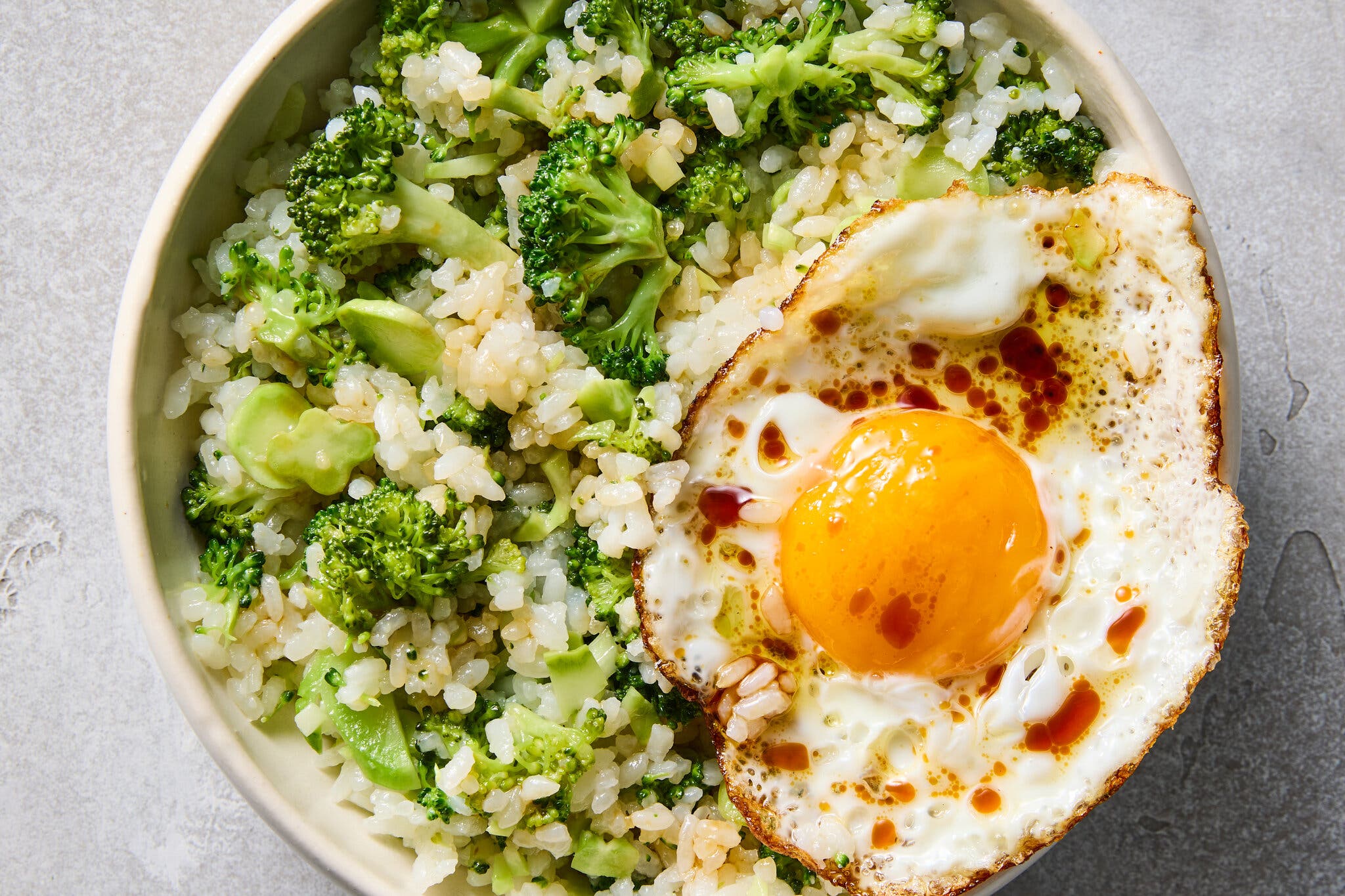 A tight overhead shot of a bowl of light-colored rice and chopped broccoli, topped with a fried egg with a bright yellow yolk.