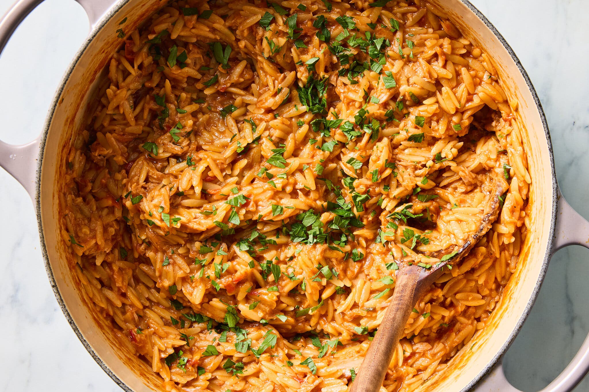 A tight overhead shot of orzo pasta cooked in a rich, red-orange sauce, topped with chopped parsley in a white pot with a wooden spoon.