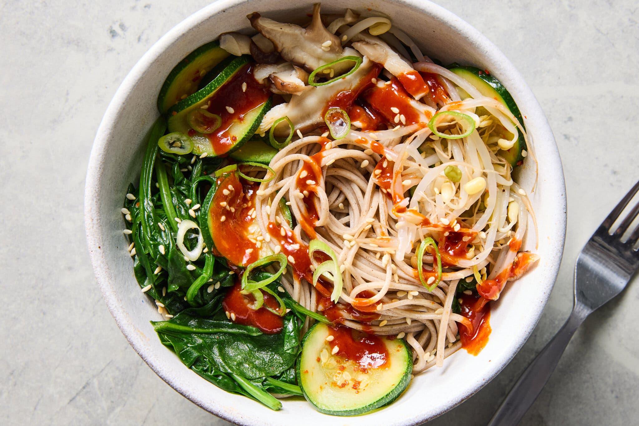 A white bowl of soba noodles topped with spinach, zucchini slices, bean sprouts, mushrooms, sliced scallions and hot sauce.