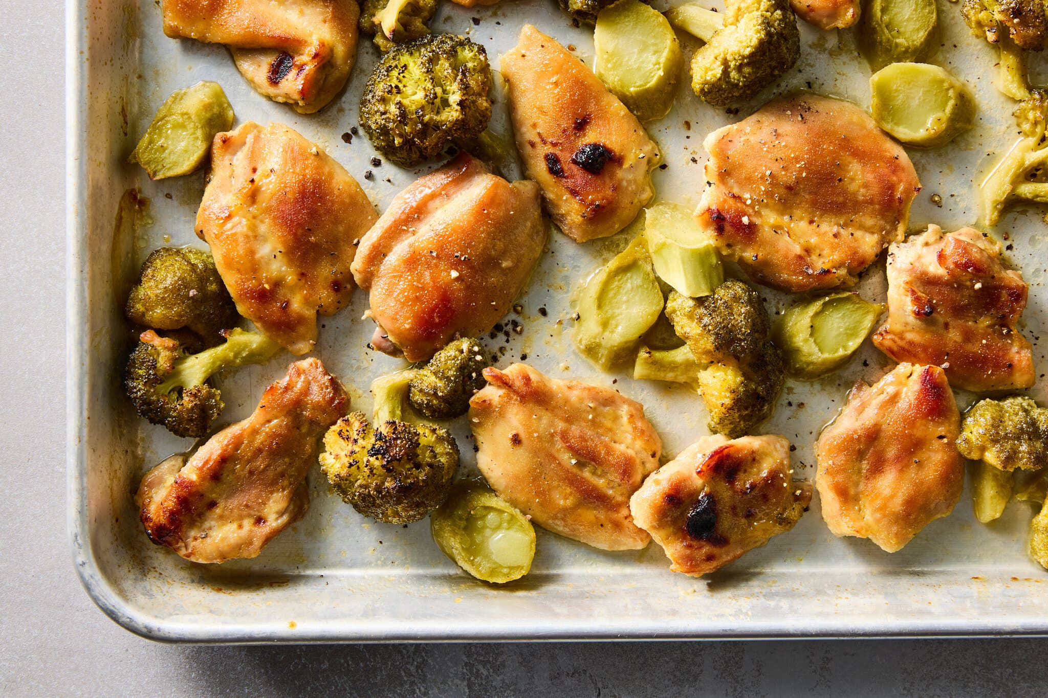 A tight overhead image of golden-brown baked chicken thighs and florets of roasted broccoli on a sheet pan.