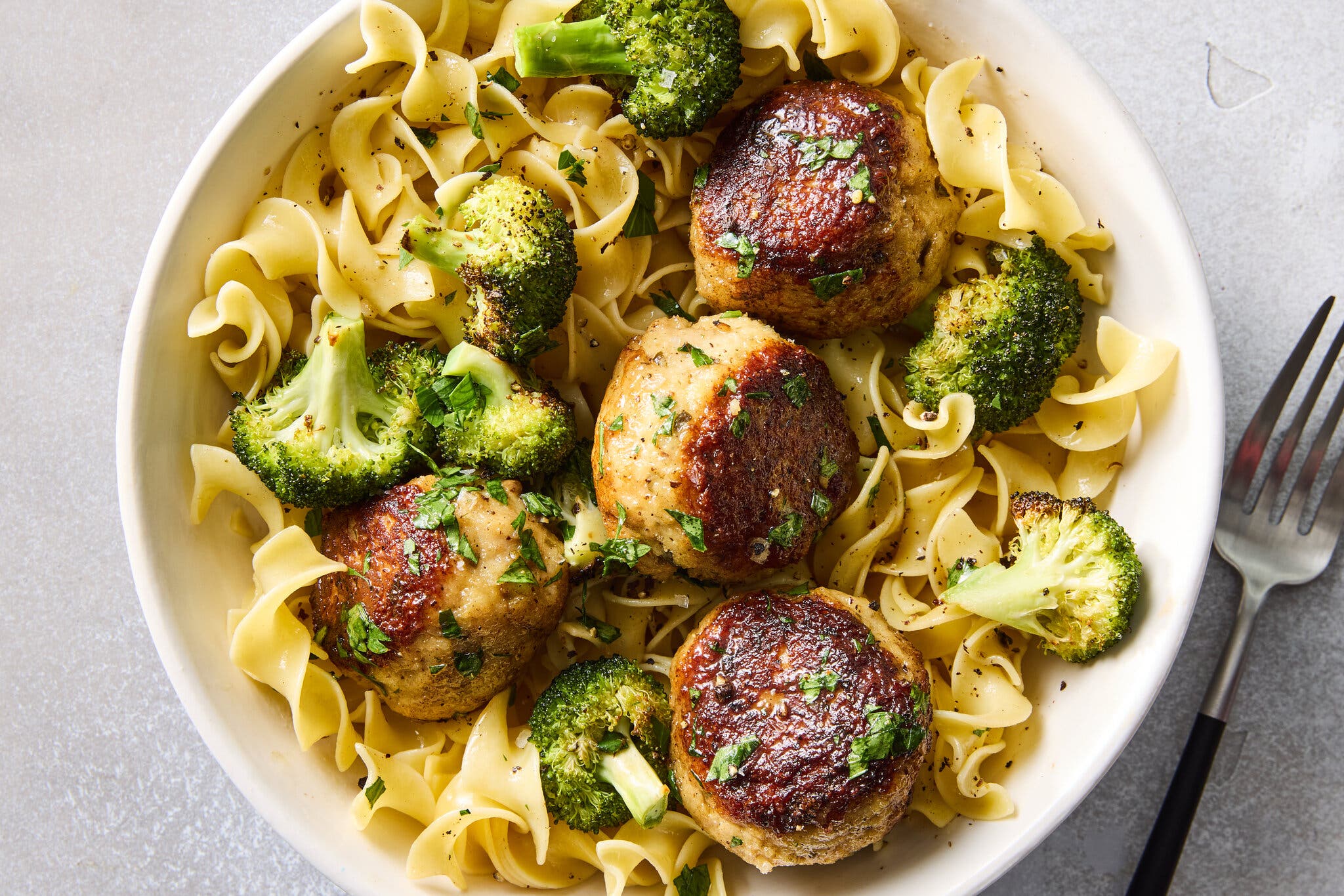 A tight overhead image of a white bowl of curly egg noodles topped with five meatballs, roasted broccoli florets and chopped parsley.