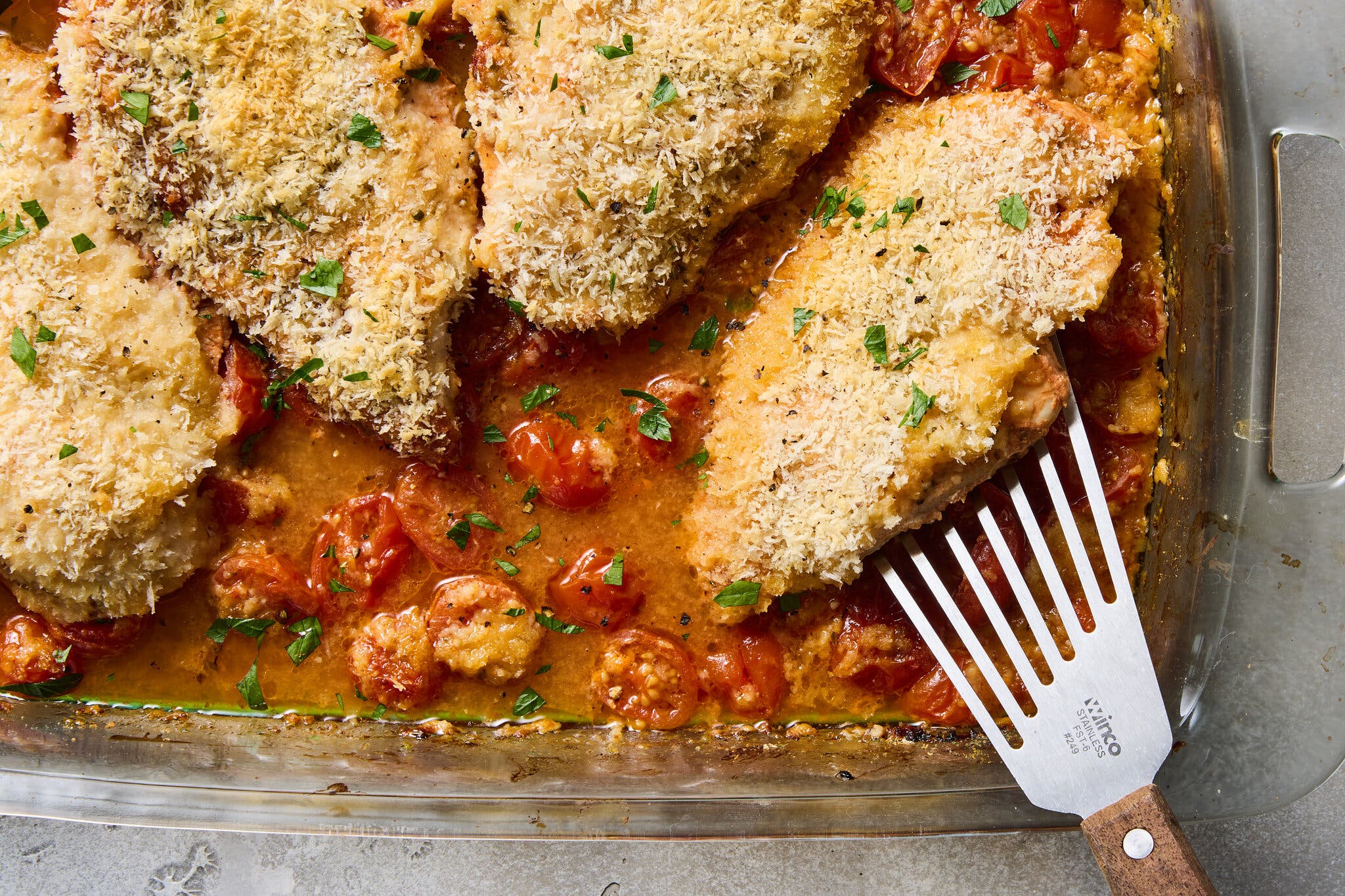 A tight overhead image of breaded chicken cutlets with parsley on a bed of roasted cherry tomatoes and sauce in a clear glass baking dish; a metal spatula lifts one piece.