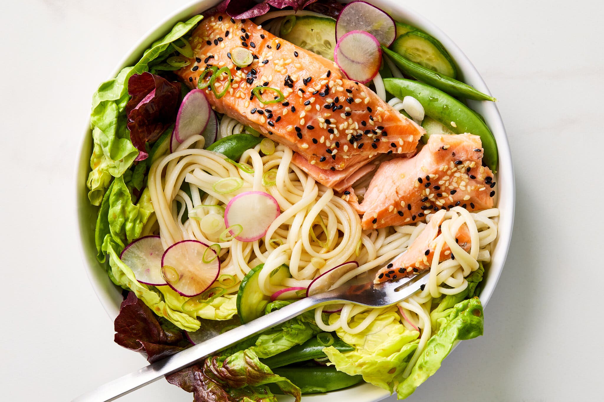 A tight overhead image of a white bowl of noodles and greens topped with sesame seed-crusted salmon, sliced cucumbers and radishes, and snow peas.