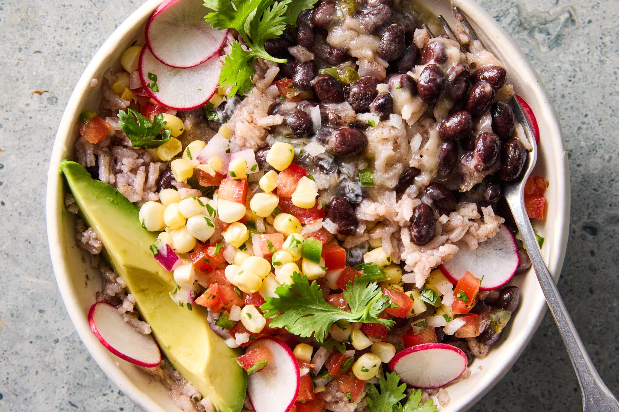 A tight overhead image of a white bowl of rice and black beans topped with corn, diced tomato, sliced radish, fresh cilantro and a large slice of avocado.