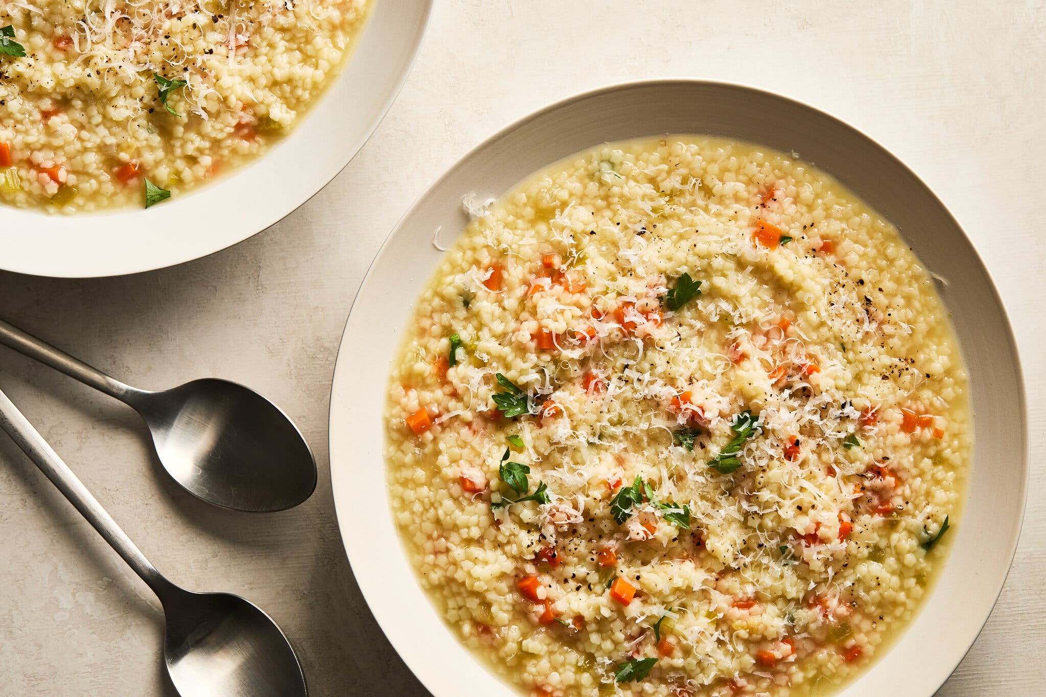 A white bowl of brothy soup with tiny pasta, carrots, celery and parsley, topped with grated Parmesan; two spoons are in the foreground.