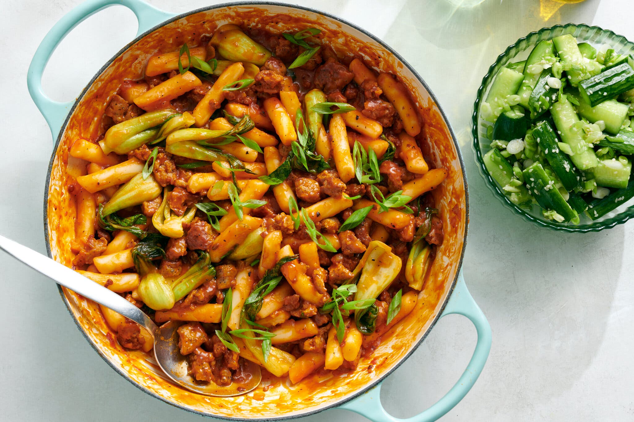 A pale blue pot of tteokbokki, Korean rice cakes, ground sausage and wilted spinach in a red-orange sauce, garnished with scallions; a side of chopped cucumber is visible to the right.