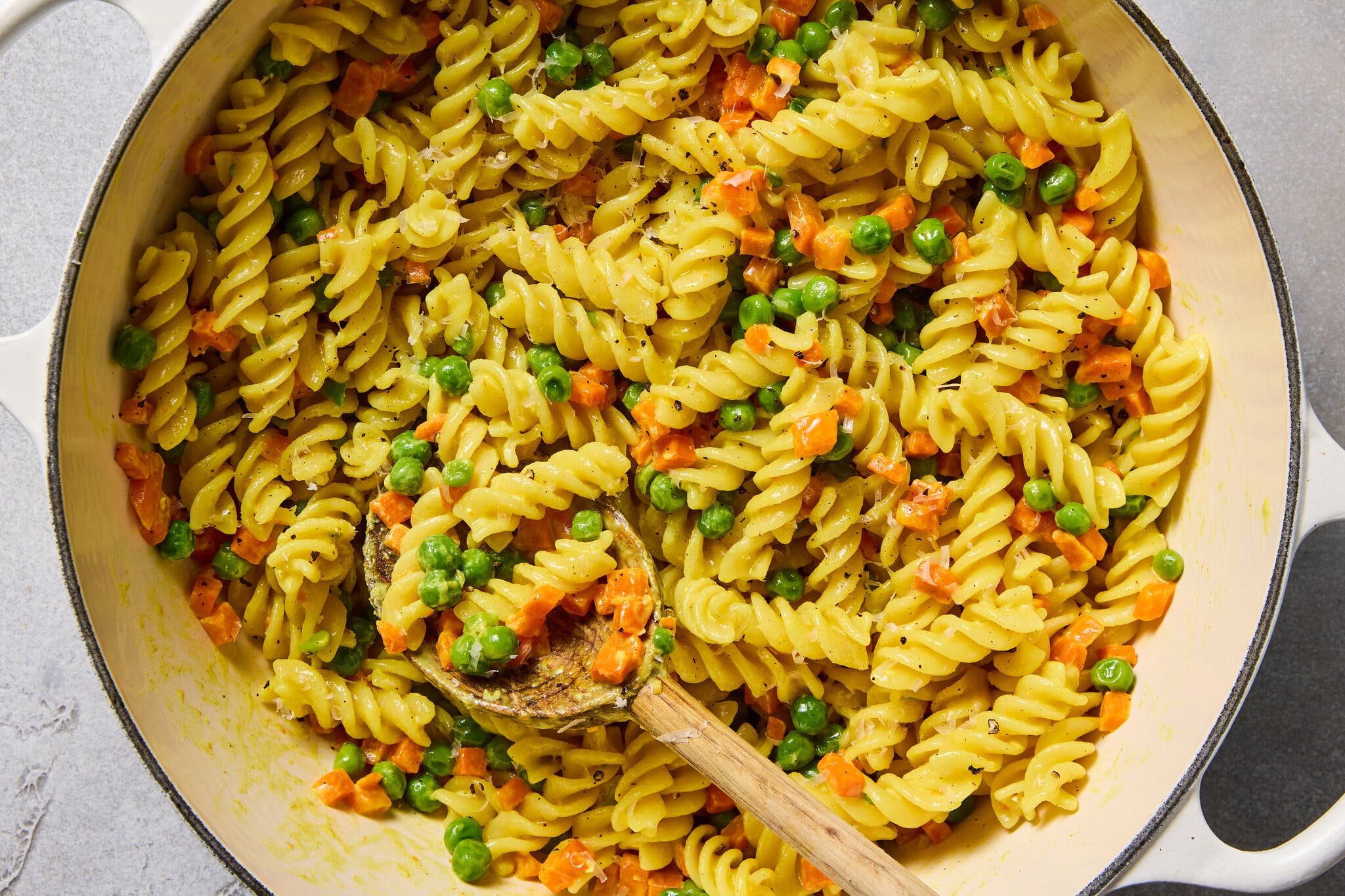 A close-up of fusilli pasta tossed with bright green peas and diced orange carrots in a white enamel cast-iron pot.
