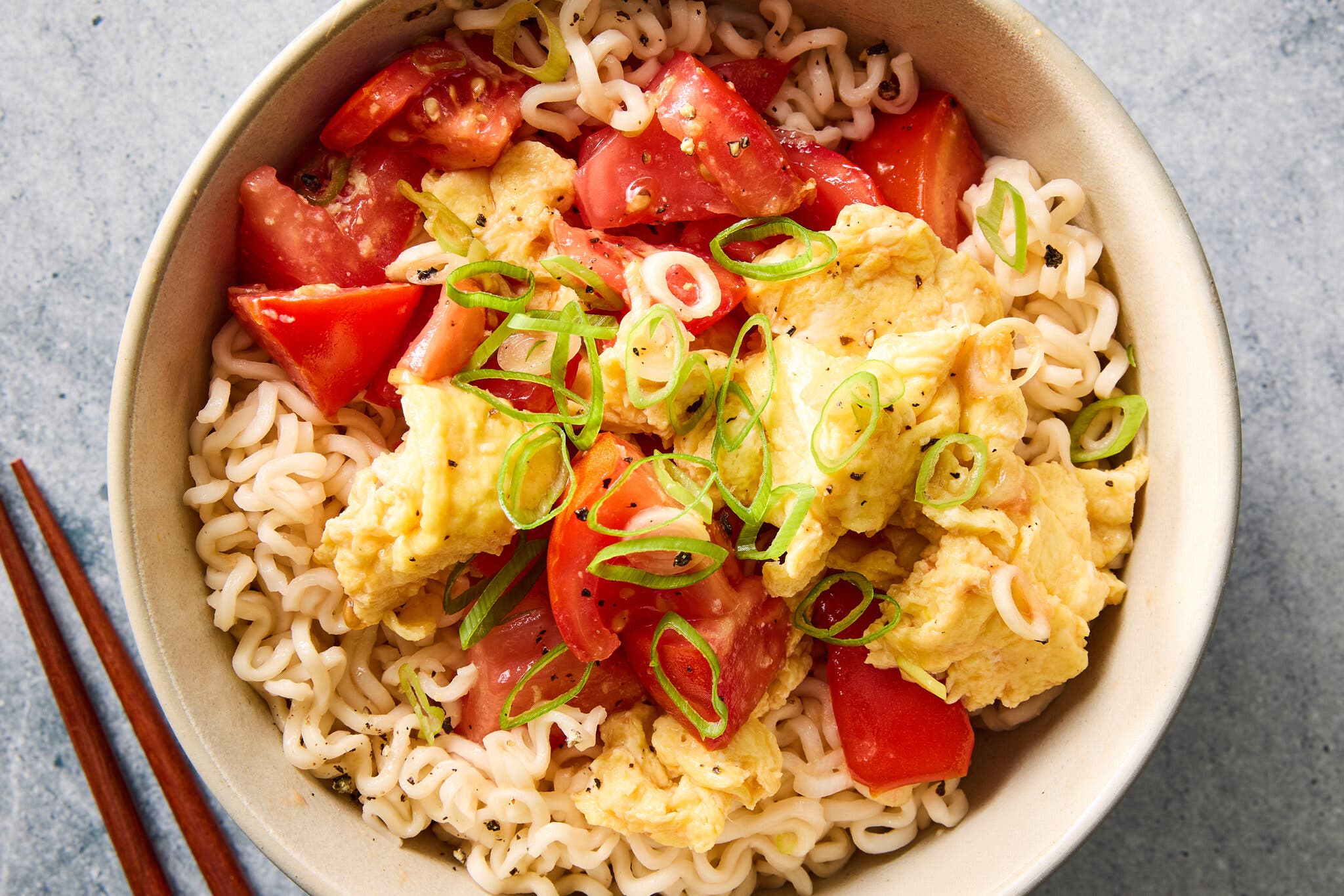A tight overhead image of a white bowl of ramen noodles topped with chunks of red tomato, yellow scrambled eggs and sliced green scallions.