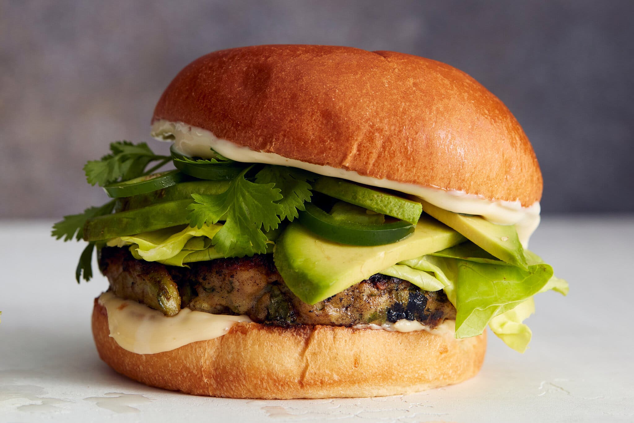 A chicken burger on a hamburger bun slicked with a creamy sauce, with lettuce, sliced avocado, jalapeños and fresh cilantro leaves.