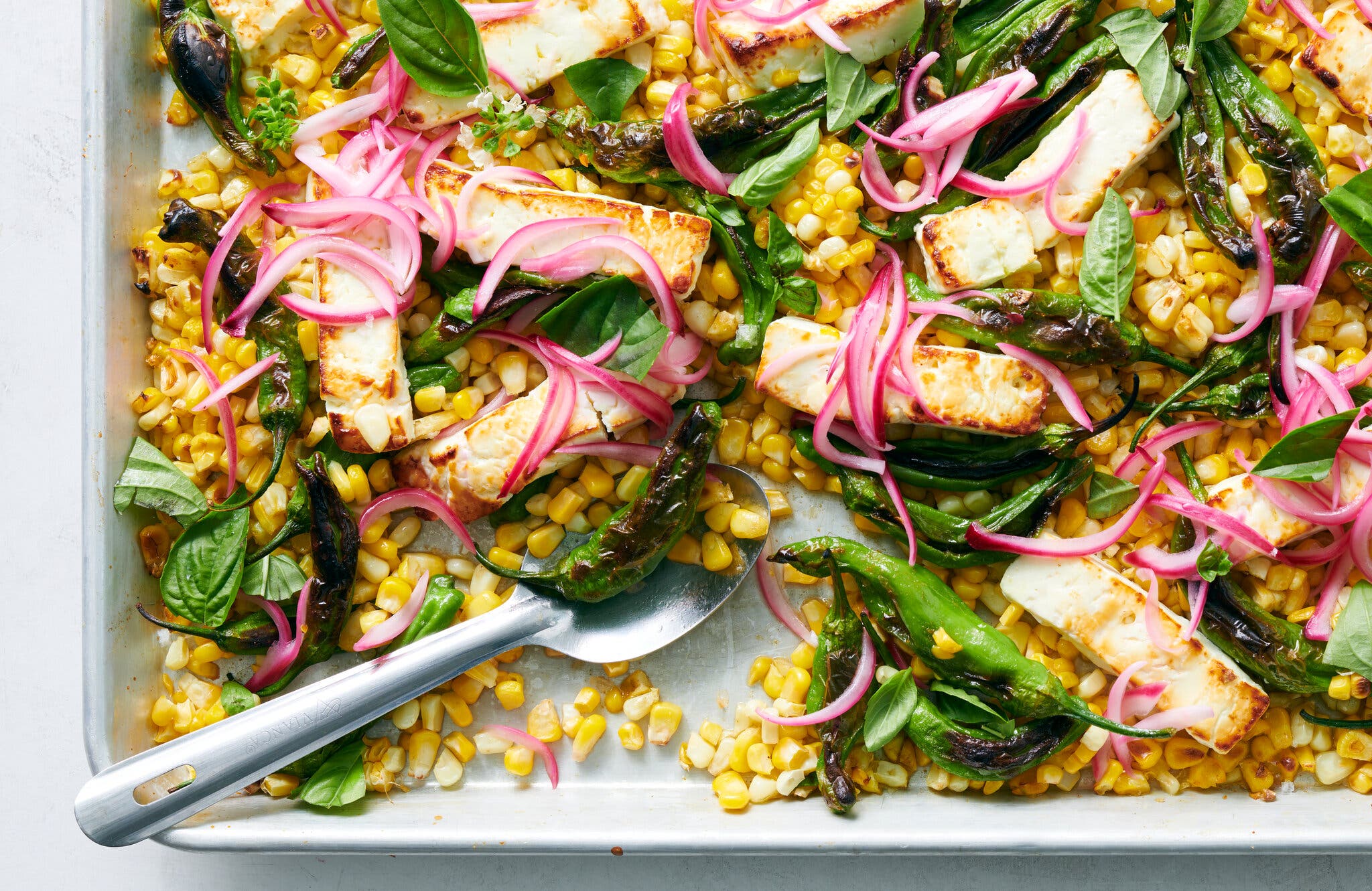An overhead image of a sheet pan filled with blistered shishitos, roasted yellow corn, feta cheese slices, fresh basil, and bright pink pickled red onions.