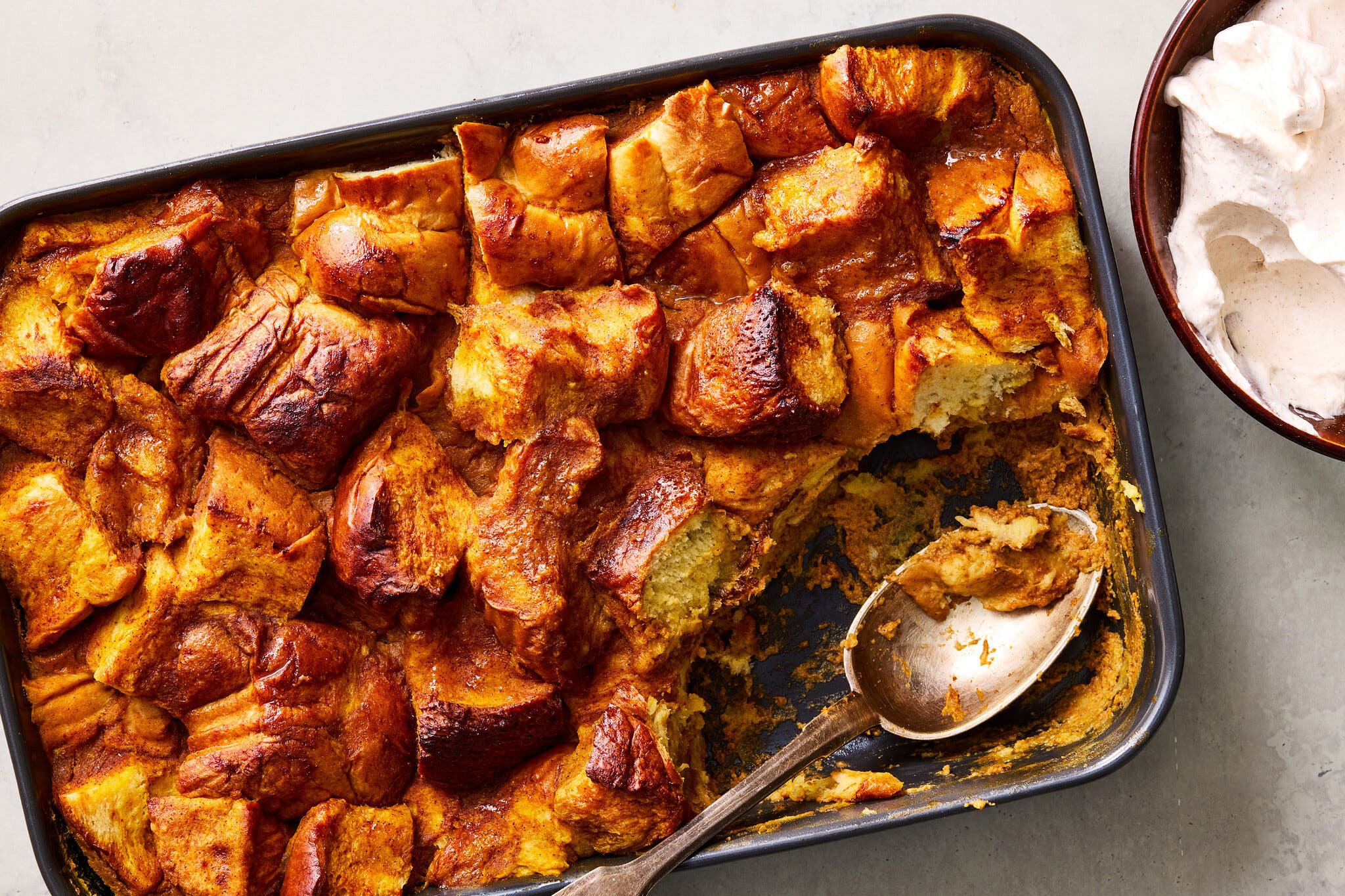 An overhead image of pumpkin bread pudding with a scoop missing. To the top right of the frame, there’s a bowl of whipped cream.