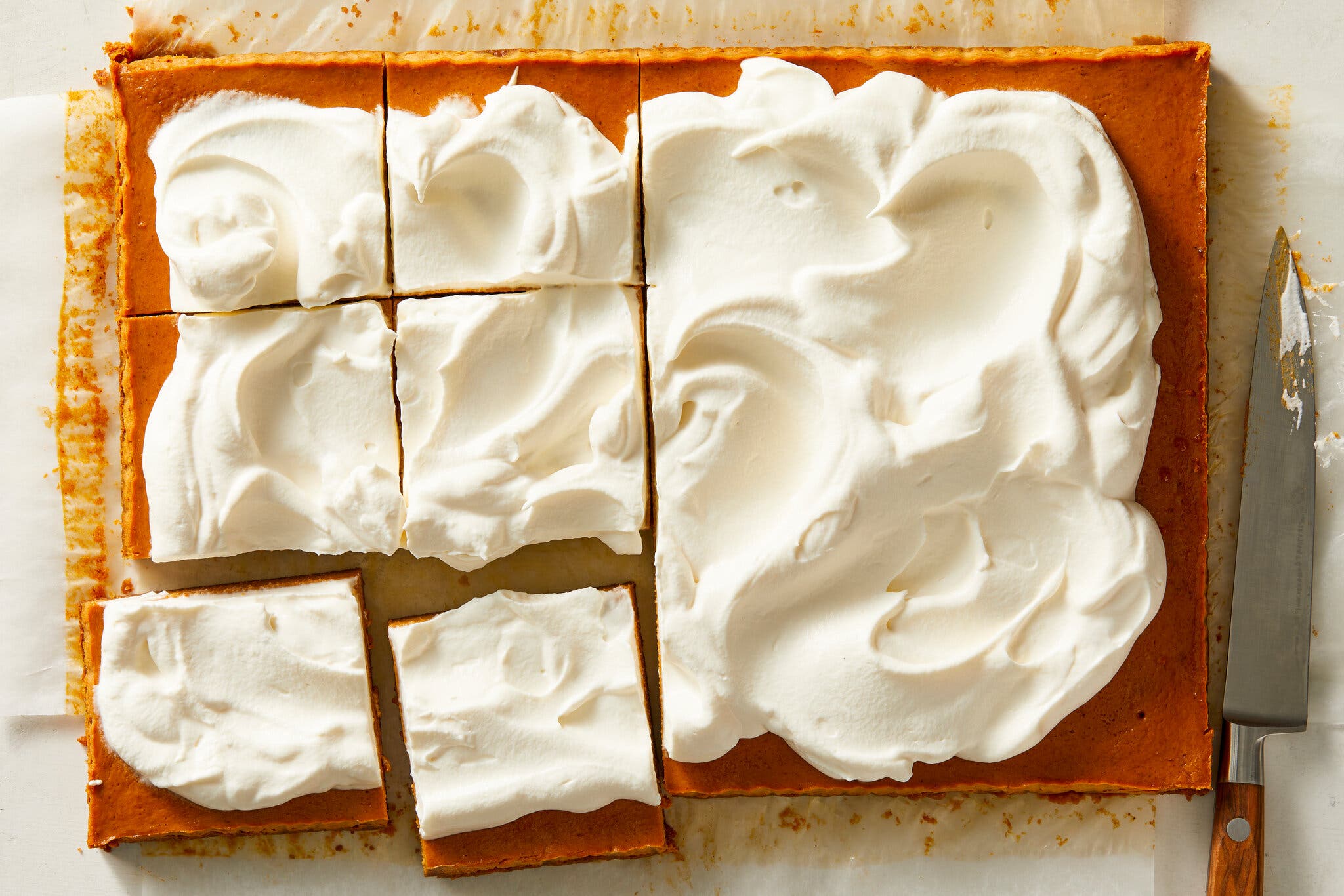 An overhead image of pumpkin pie bars topped with whipped cream on parchment paper. Six square slices have been made. To the right there is a knife.