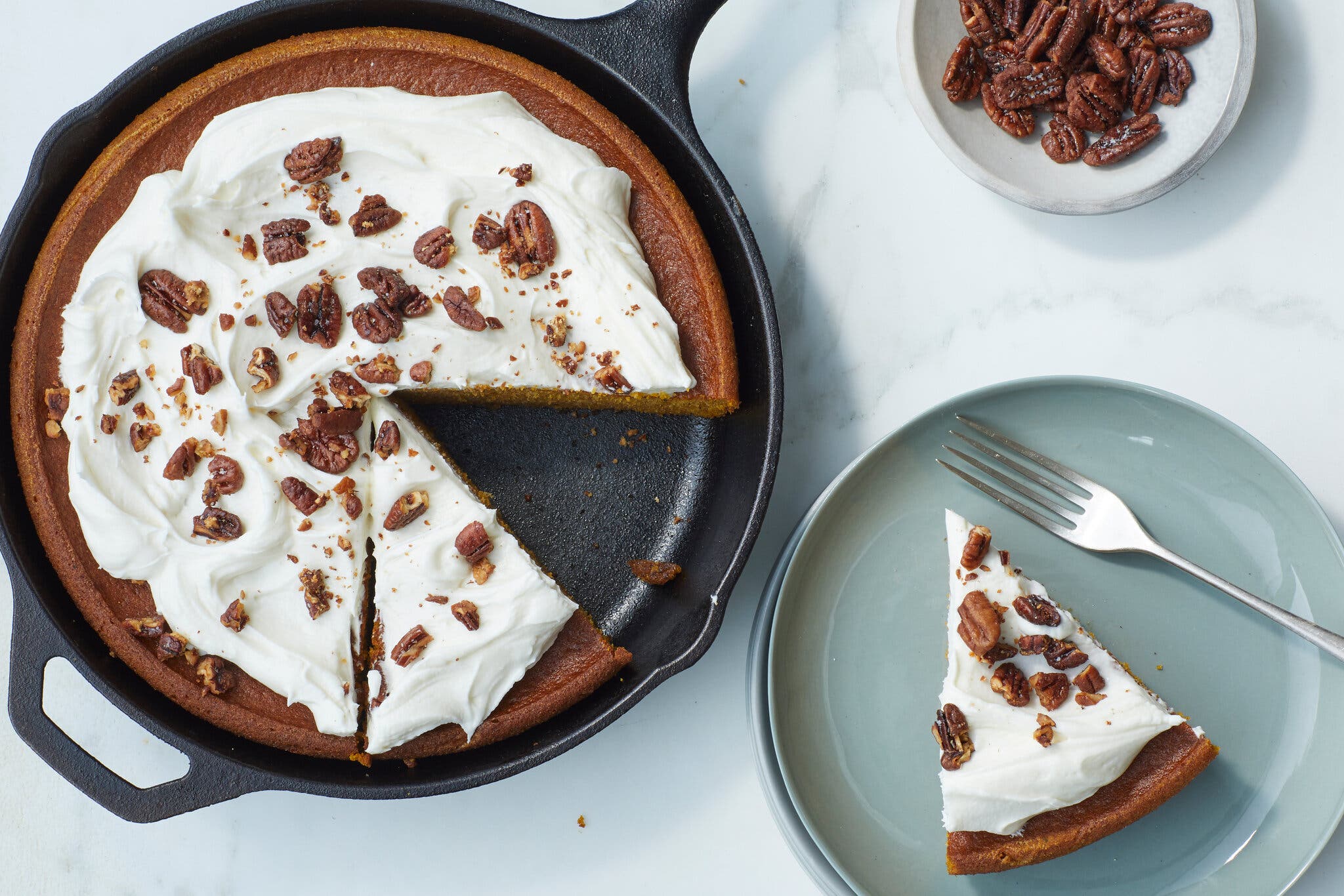 An overhead image of a pumpkin pie skillet cake topped with cream cheese frosting and chopped pecans. In the bottom-right corner of the frame, there is a blue plate with a slice of cake and a fork.
