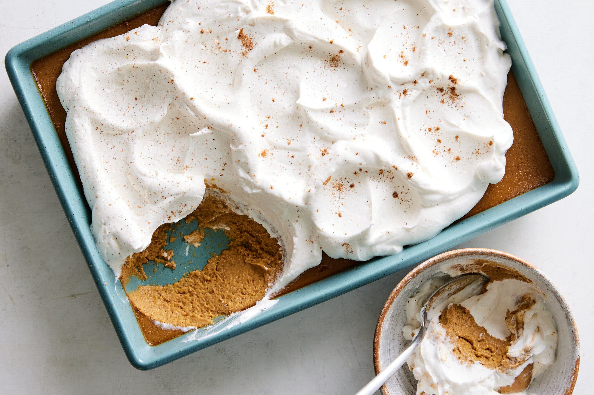 An overhead image of a blue baking dish holds a light-brown custard topped with whipped cream that’s been sprinkled with a brown spice. A serving sits in a bowl to the bottom right.