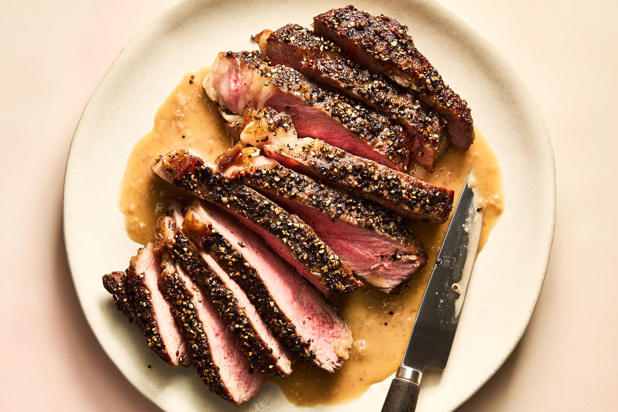 An overhead image of a plate of a sliced rib-eye steak crusted in crushed peppercorns on a bed of peppery, Cognac pan sauce.