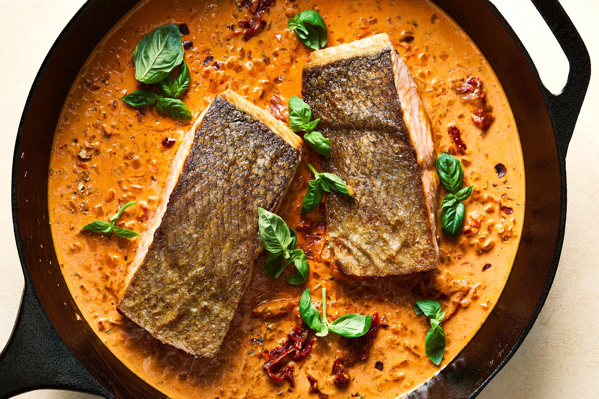 An overhead image of a cast iron skillet with two salmon fillets, skin-side up, in a creamy sun-dried tomato sauce topped with fresh basil.