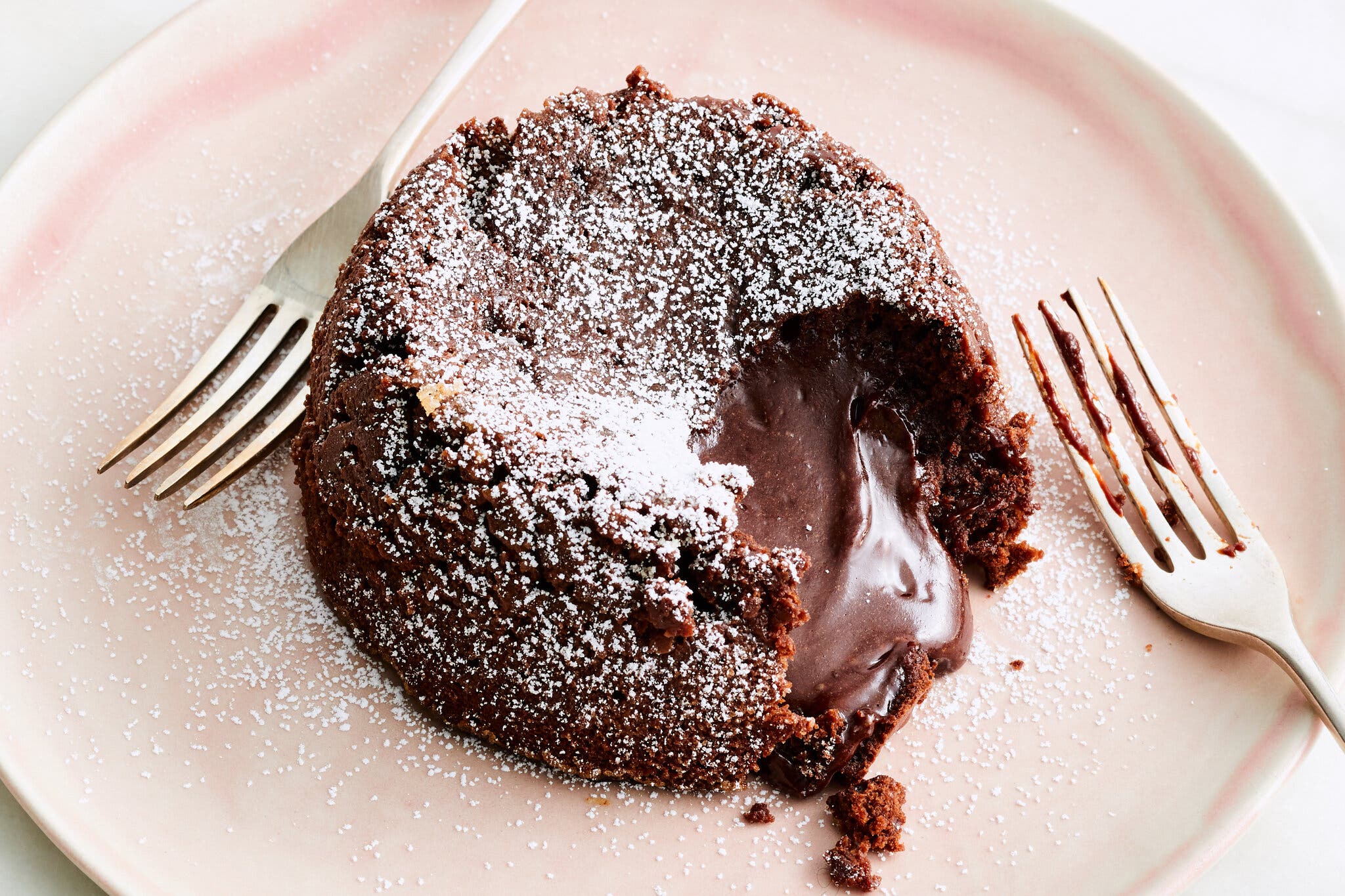 A chocolate lava cake on a pink plate with two forks. A bite of the cake is missing, revealing the melted chocolate sauce inside.