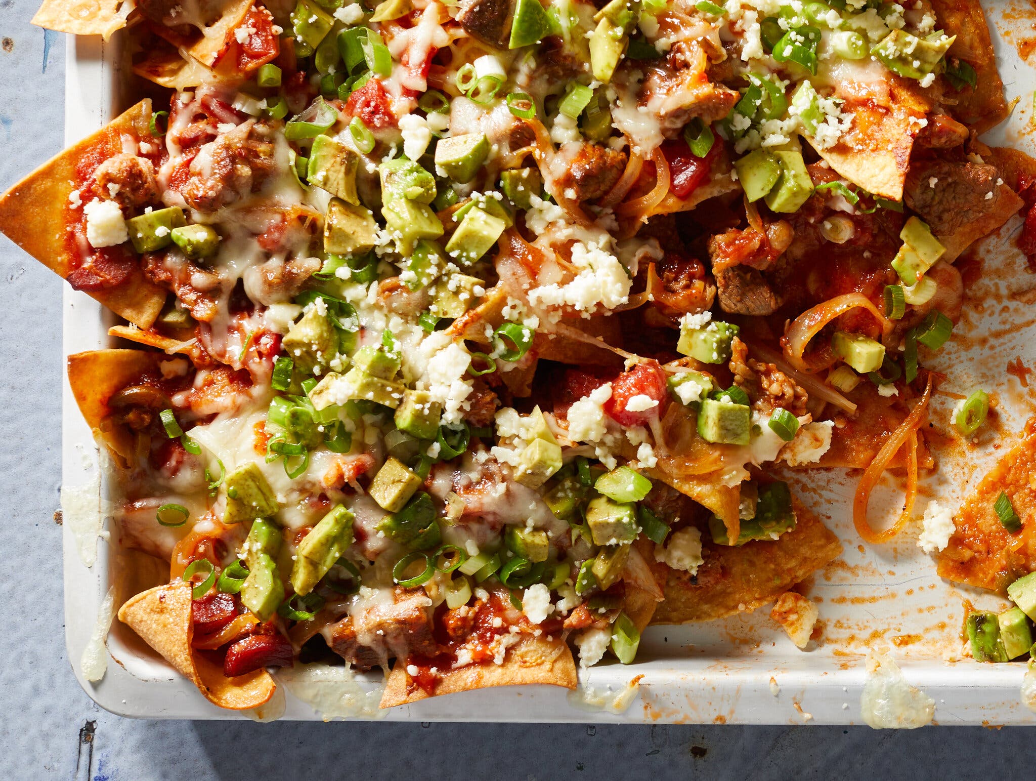 An overhead image of a pan of cheesy bricklayer-style nachos topped with avocado, queso fresco and scallions.