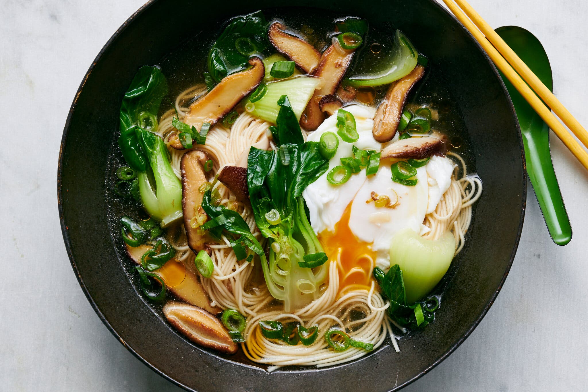 An overhead image of a bowl of somen noodle soup with mushrooms, bok choy, a poached egg and scallions.