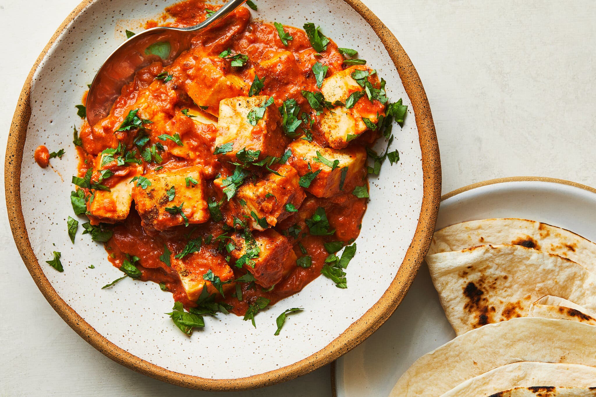 An overhead image of a bowl of butter paneer. In the bottom right corner there’s a plate of roti.