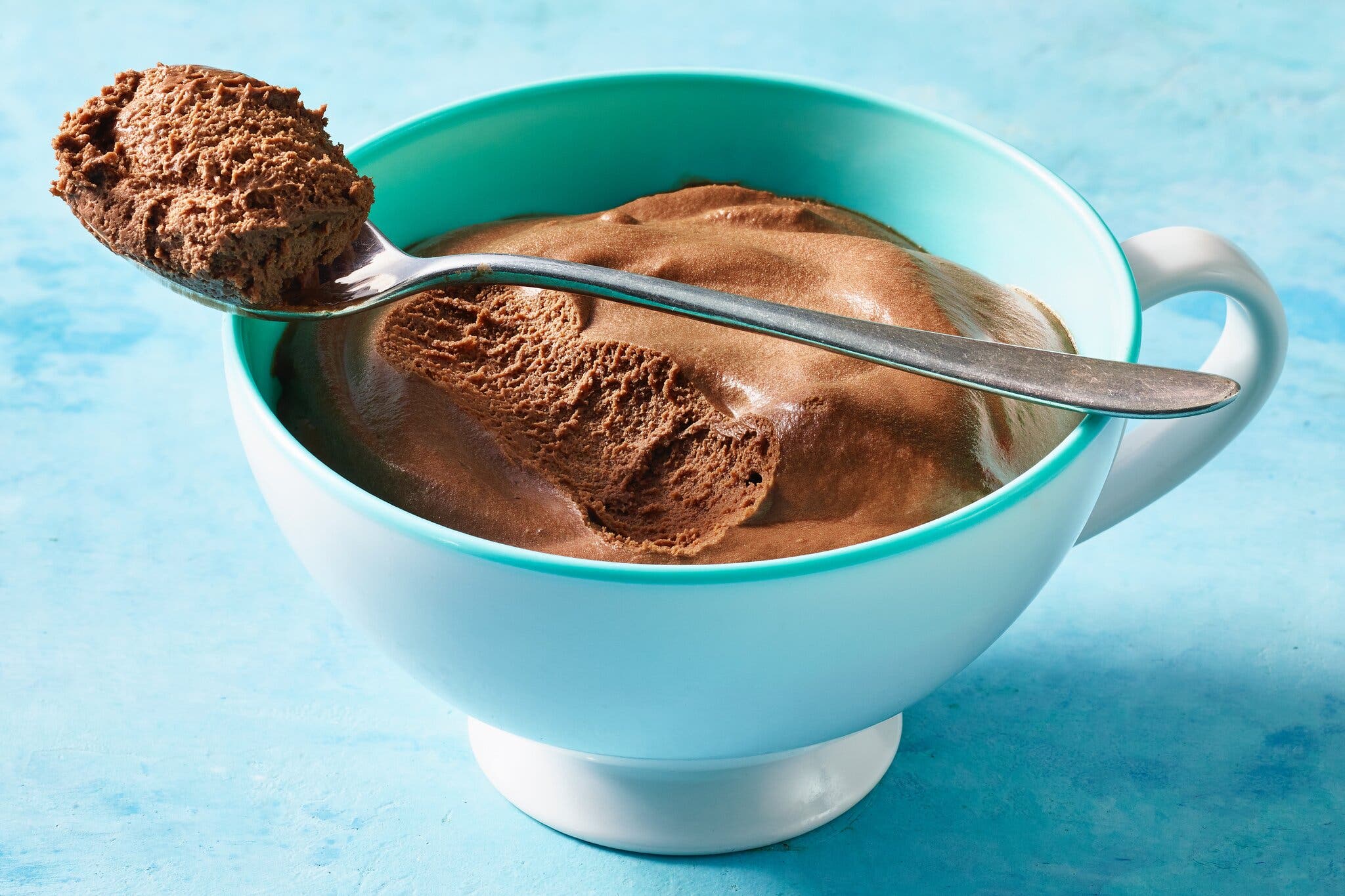 An aqua cup holds a serving of chocolate mousse. Atop the cup is a spoon holding a bit of mousse.