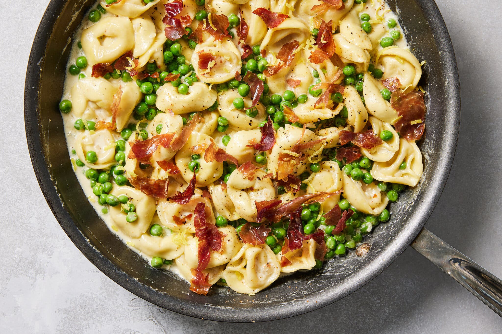An overhead image of tortellini with prosciutto and peas in a lemon cream sauce in a black pan.