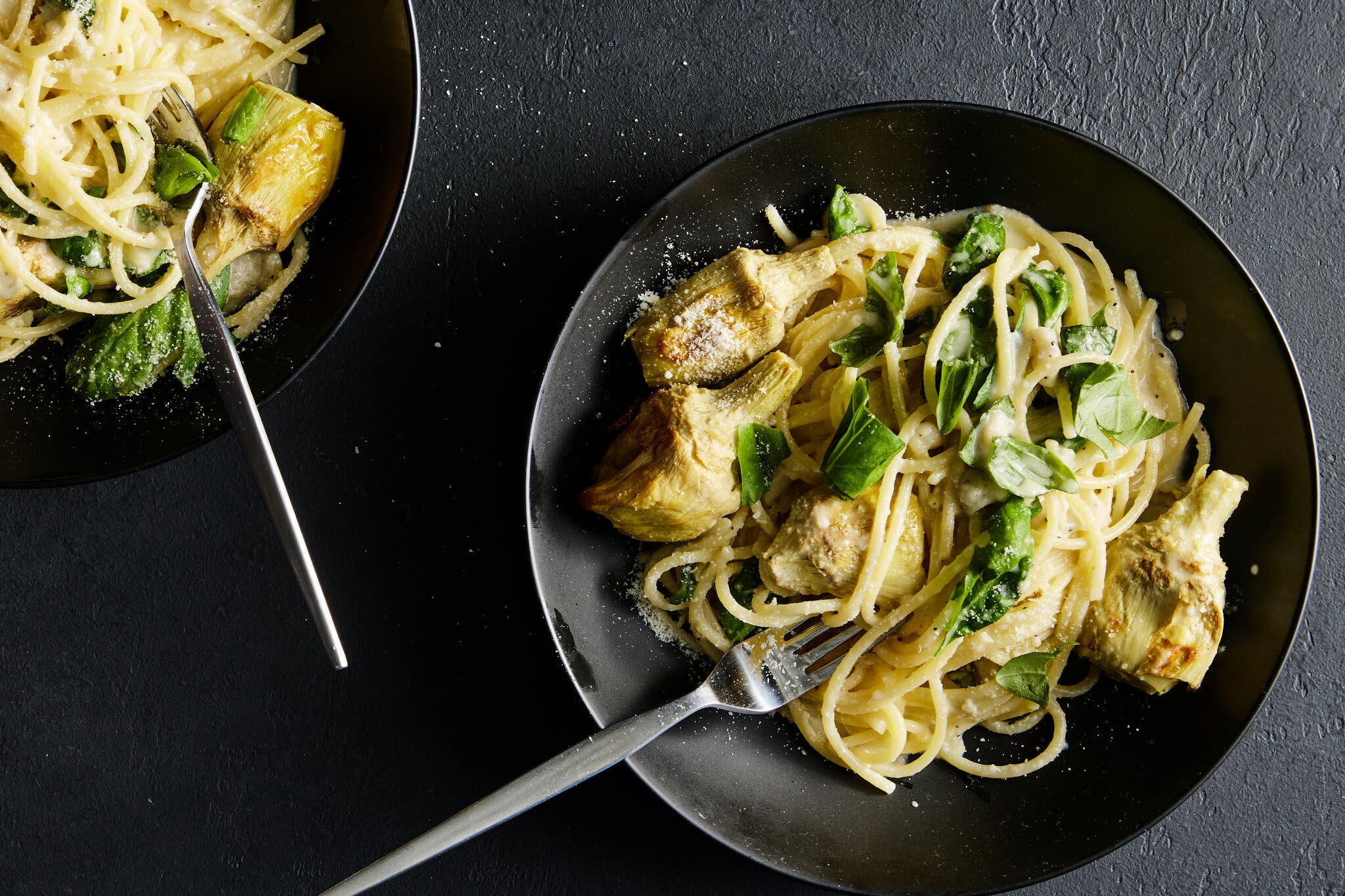 An overhead image of two black bowls with lemon spaghetti with roasted artichokes and torn basil.