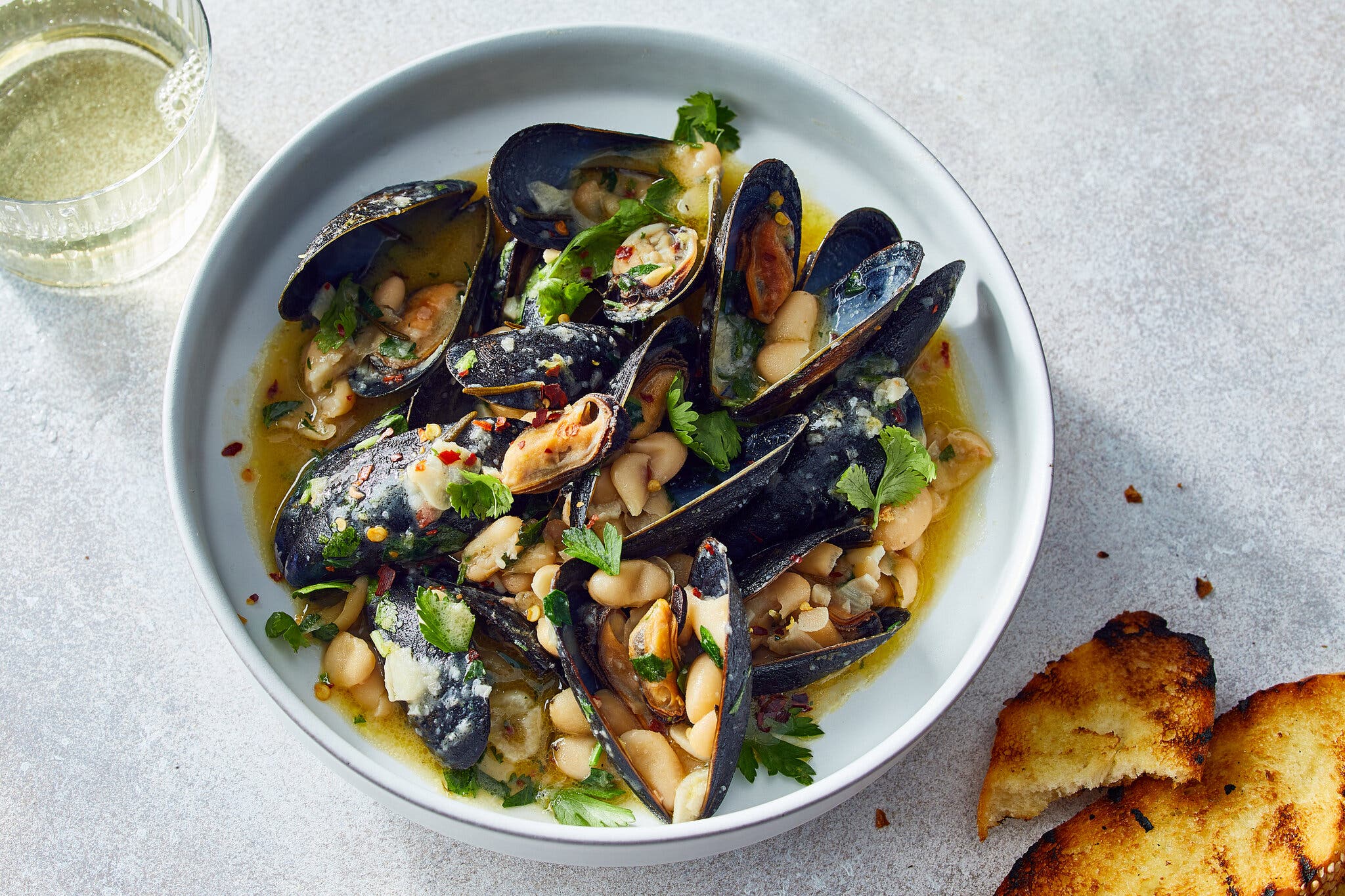 An overhead image of a bowl of mussels with white beans and garlic and pieces of toast in the bottom right corner.