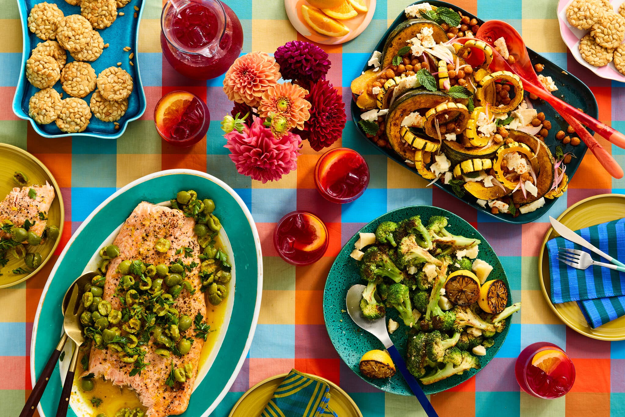 A colorful tablecloth is topped with platters of food, drink, plates and flowers.