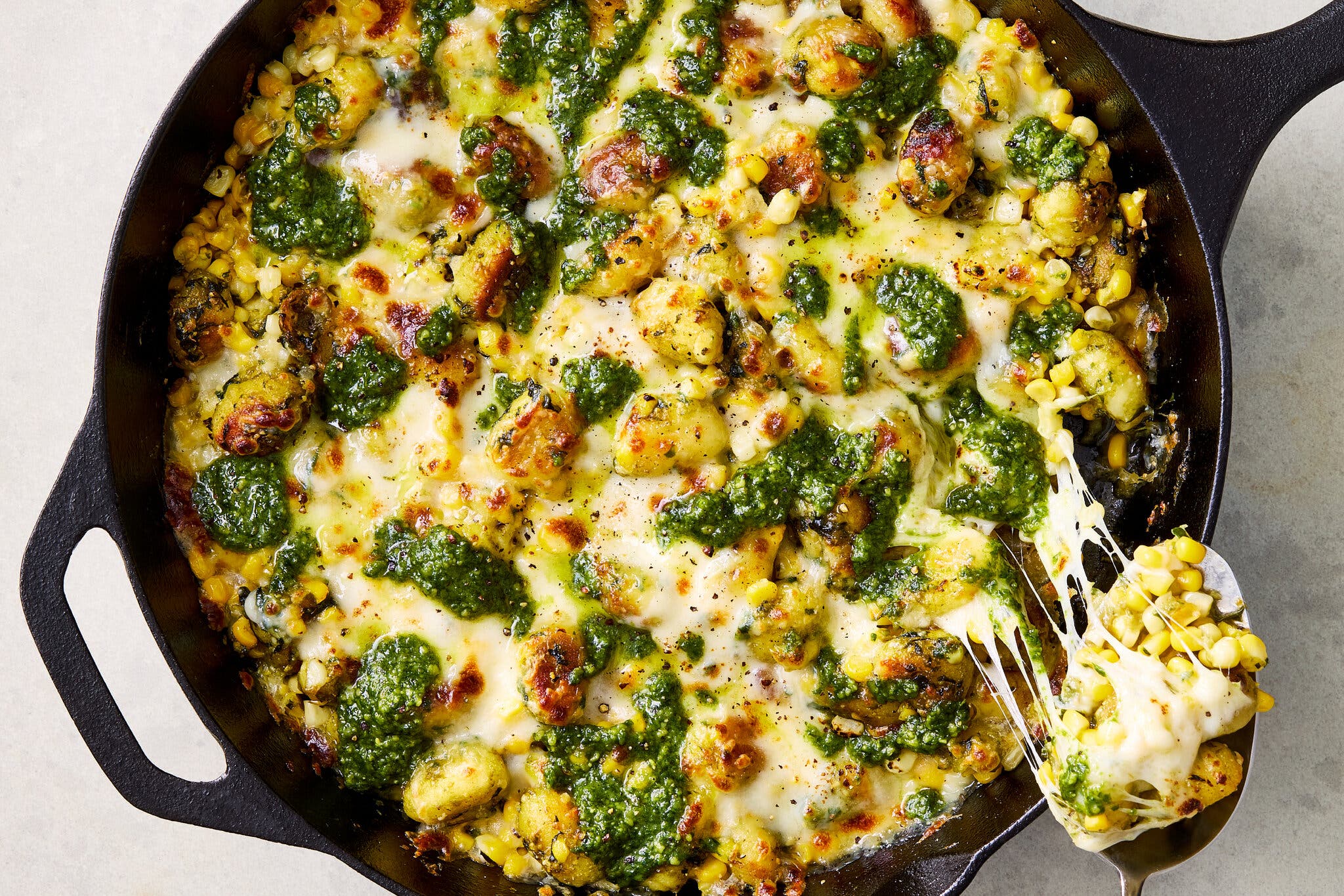 A black cast-iron skillet filled with browned gnocchi, corn kernels, melted white cheese, and dollops of bright green pesto. A silver spoon lifts a spoonful out.