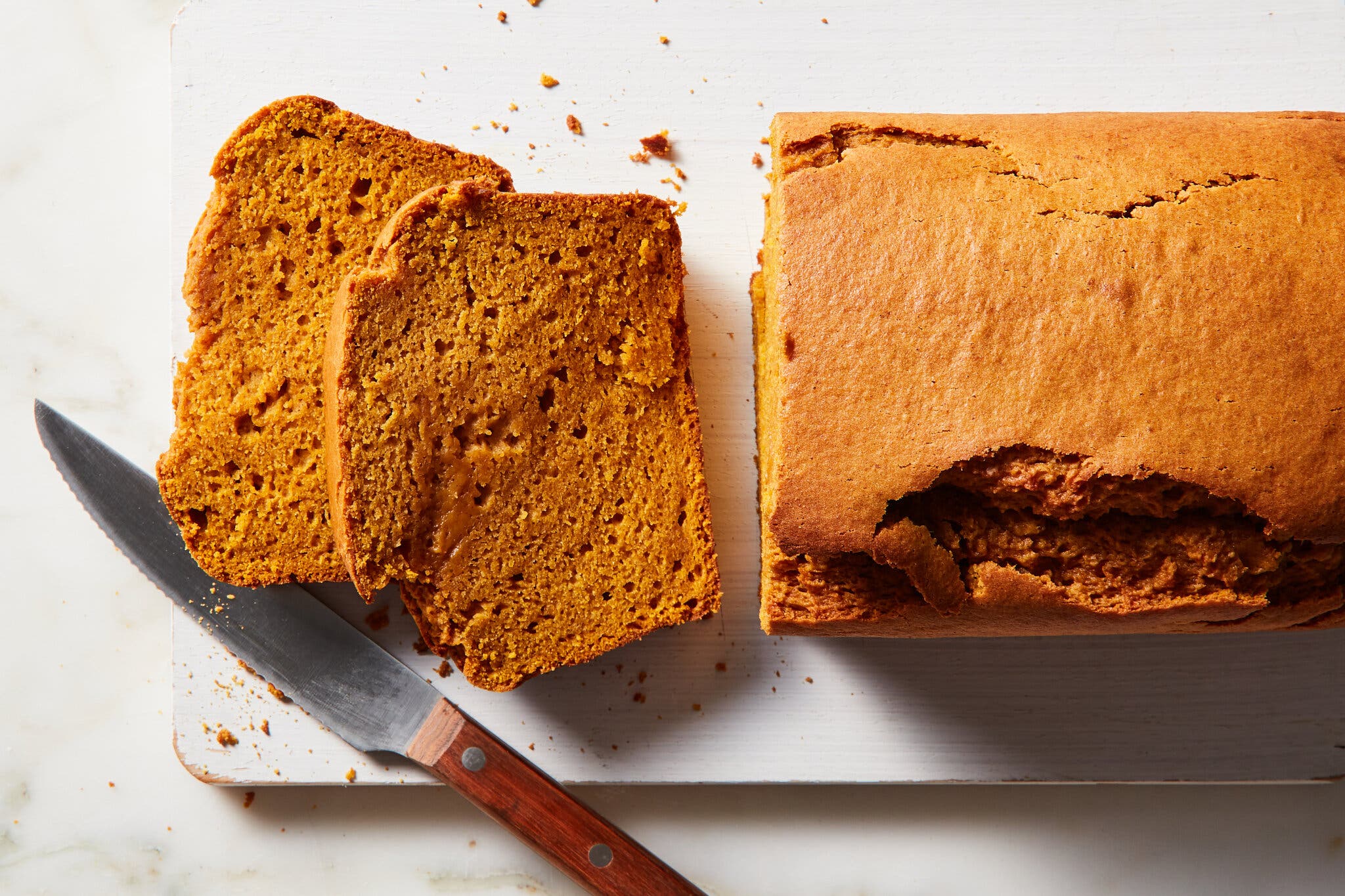 An overhead image of a loaf of pumpkin bread on a cutting board, with two slices and a knife.