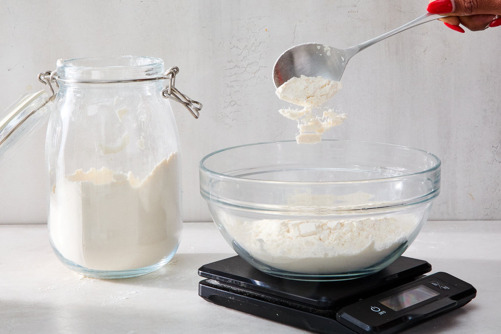 A side image of a spoon dropping flour into a bowl on a scale. A larger vessel of flour sits off to the side.