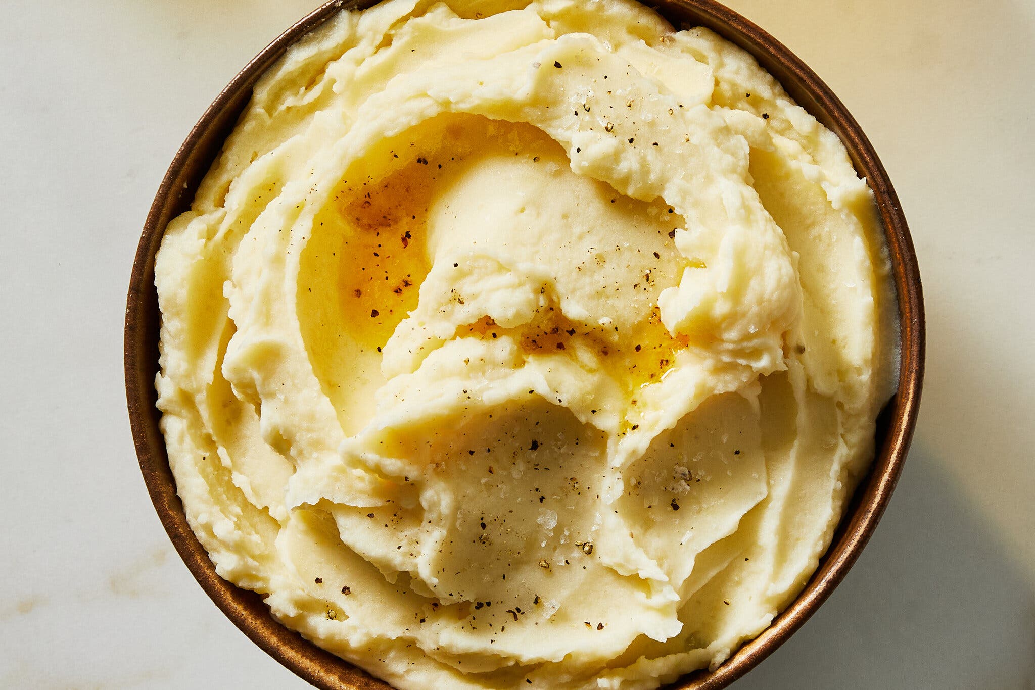 An overhead image of a bowl of mashed potatoes topped with melted butter, flaky salt and crushed black pepper.