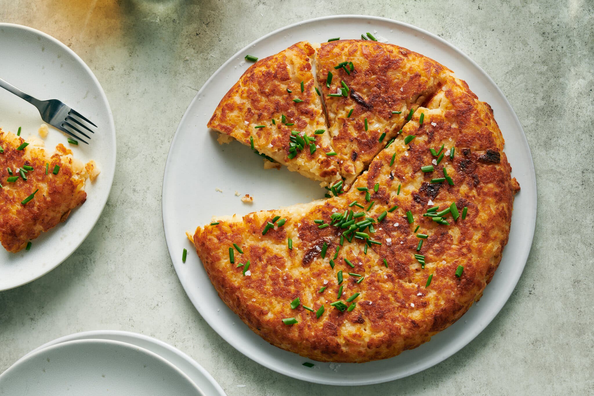 An overhead photo shows a browned mashed potato cake with a slice taken out. It sits on a white plate against a gray background. To the left is a serving with a fork on a white plate.