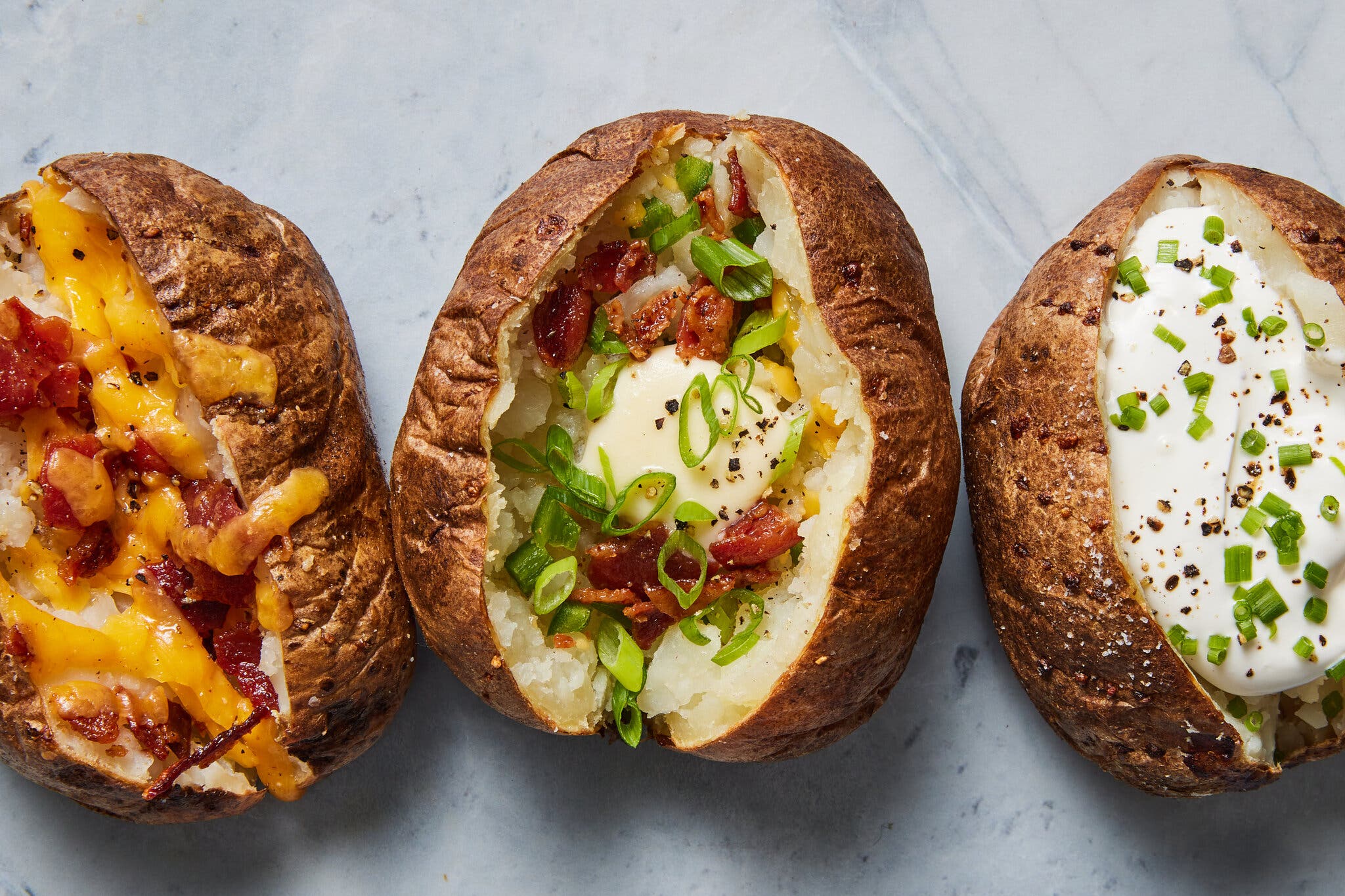 Three baked potatoes are lined up; one has Cheddar and bacon, another has butter, bacon and scallions and the third has sour cream and chives.