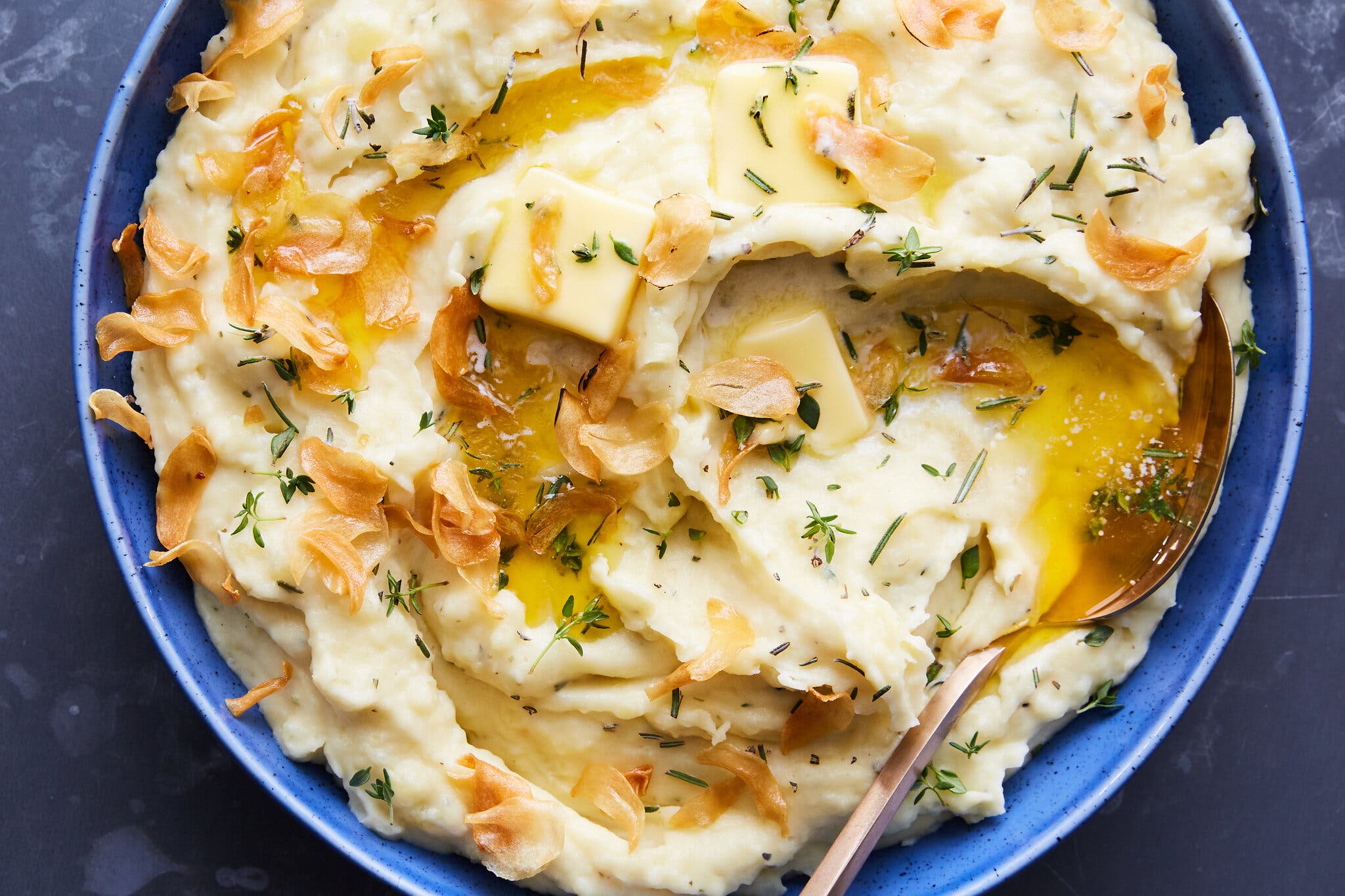 An overhead image of a blue bowl with creamy, double-garlic mashed potatoes. The dish is garnished with melted butter, garlic chips and fresh thyme. 