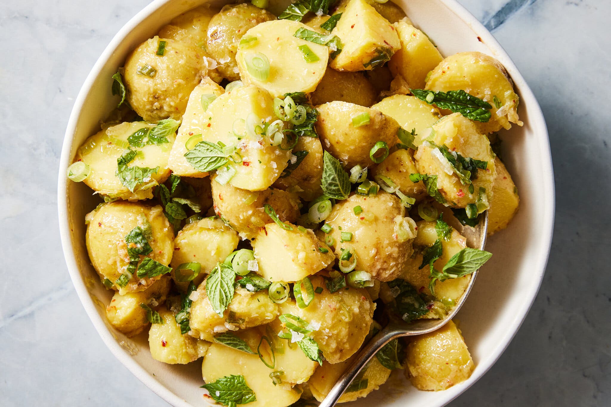 An overhead shot of lemon potato salad with mint in a white bowl with a serving spoon.