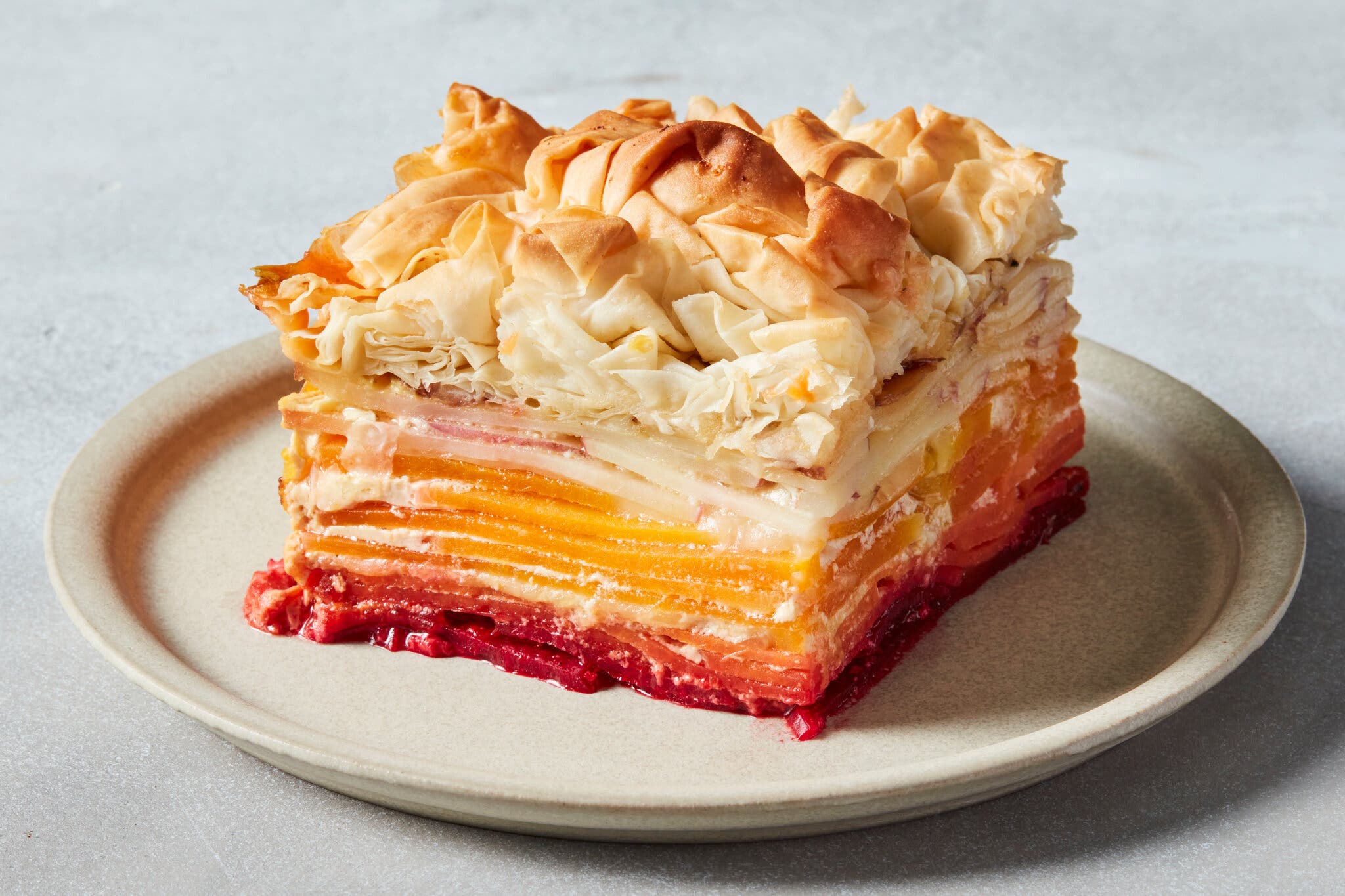 A slice of gratin — resembling a sunrise with the red of beets, the orange of squash and sweet potatoes and the rosy white of potatoes — is topped with scrunched-up phyllo.
