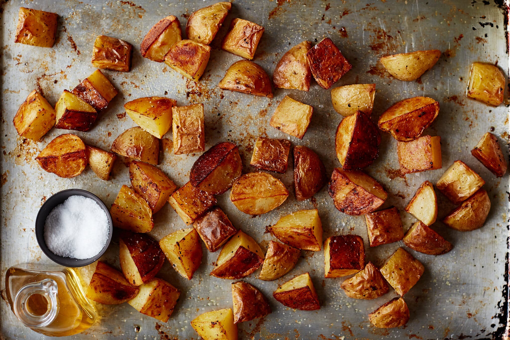 An overhead image of a sheet pan with salt and vinegar roasted potatoes. A bottle of vinegar and a pinch bowl of salt sit in the bottom left corner of the frame.