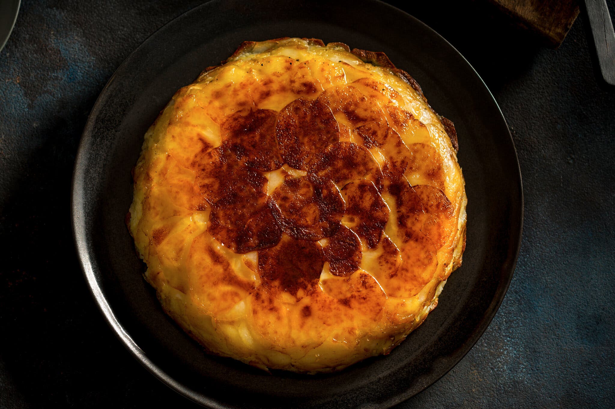 An overhead image of a burnished pommes Anna, a French potato tart on a black plate and a dark leather mat. 