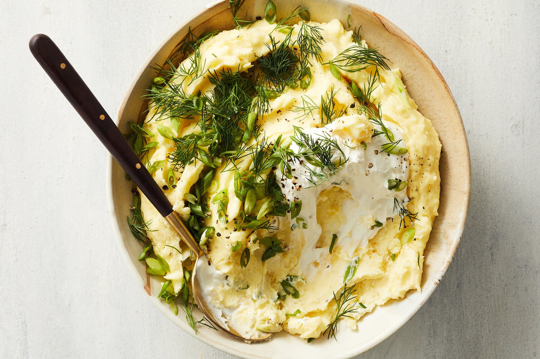 An overhead shot of a bowl of mashed potatoes topped with a dollop of labneh, scallions, dill and crushed black pepper. 
