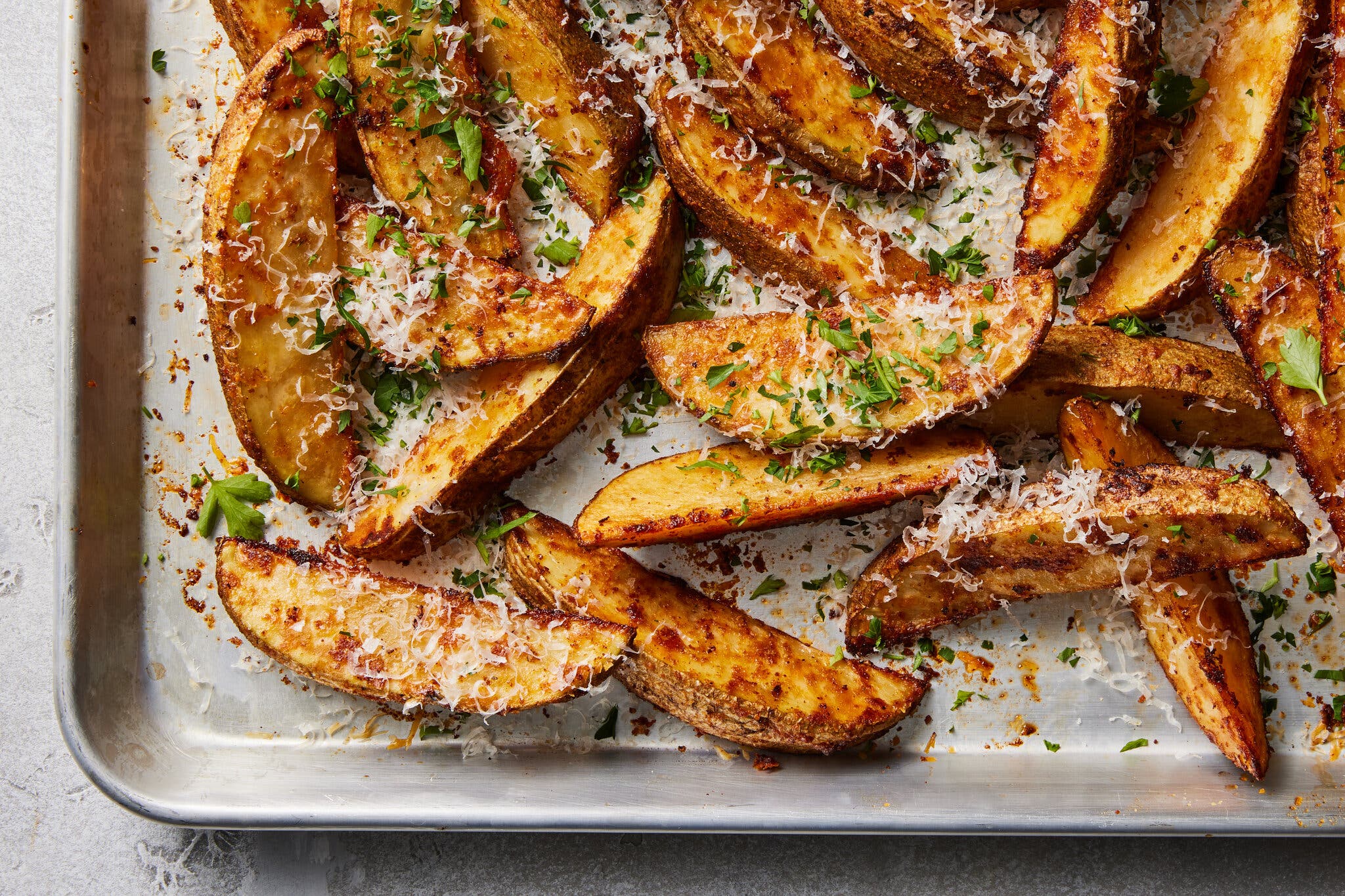 An overhead image of potato wedges on a sheet pan, garnished with grated Paremsan and chopped parsley.