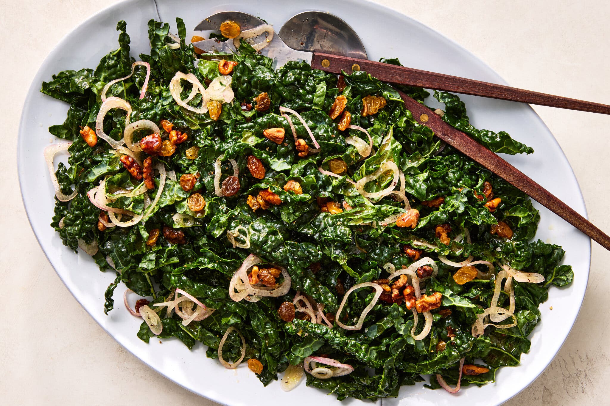 An overhead shot of a white oblong plate of kale salad with pickled raisin vinaigrette. Utensils sit on the top half of the plate.