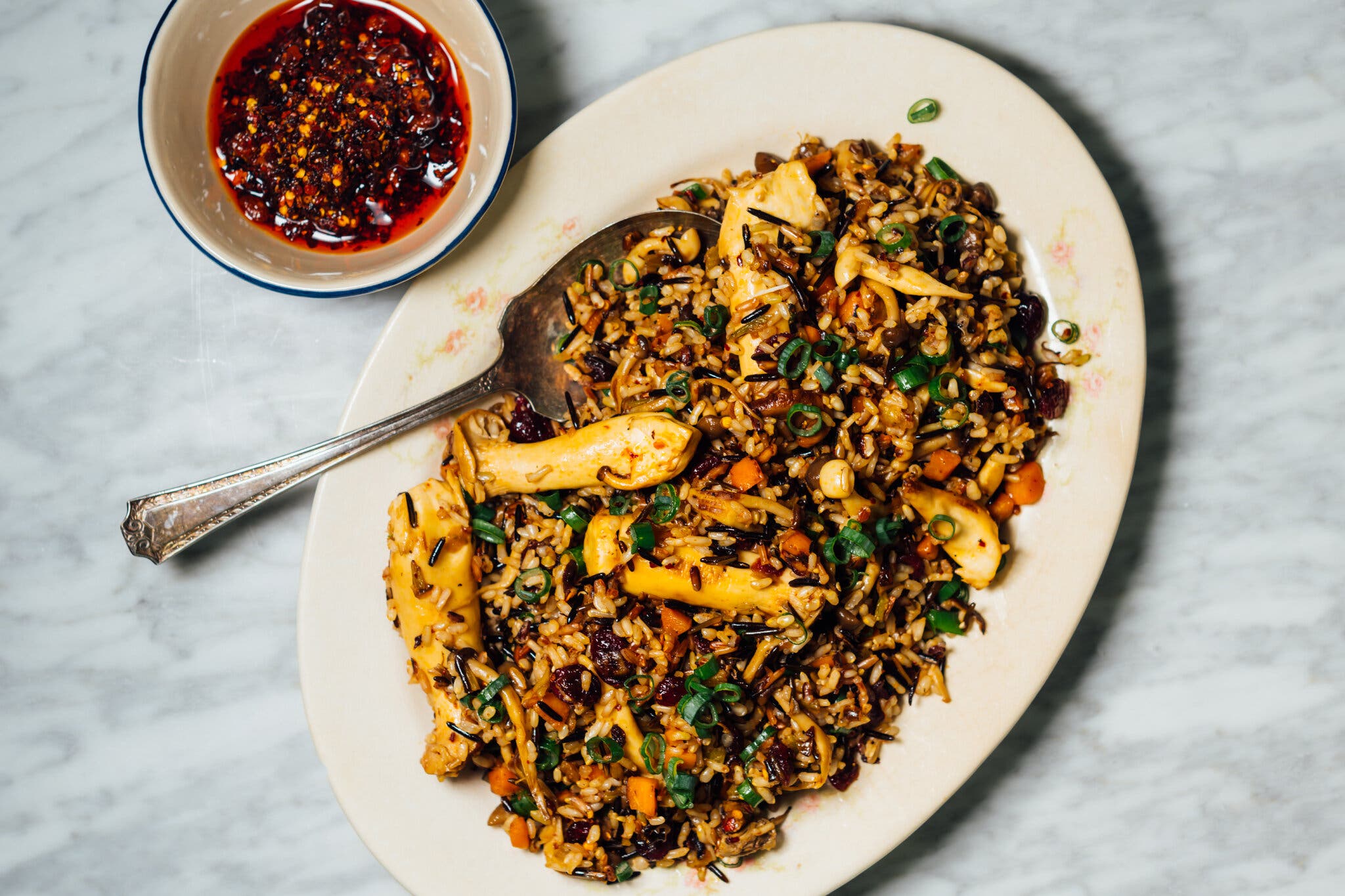 An overhead shot of a white oblong plate of wild rice stuffing and a bowl of chile crisp. A spoon juts out of the top left corner of the plate.