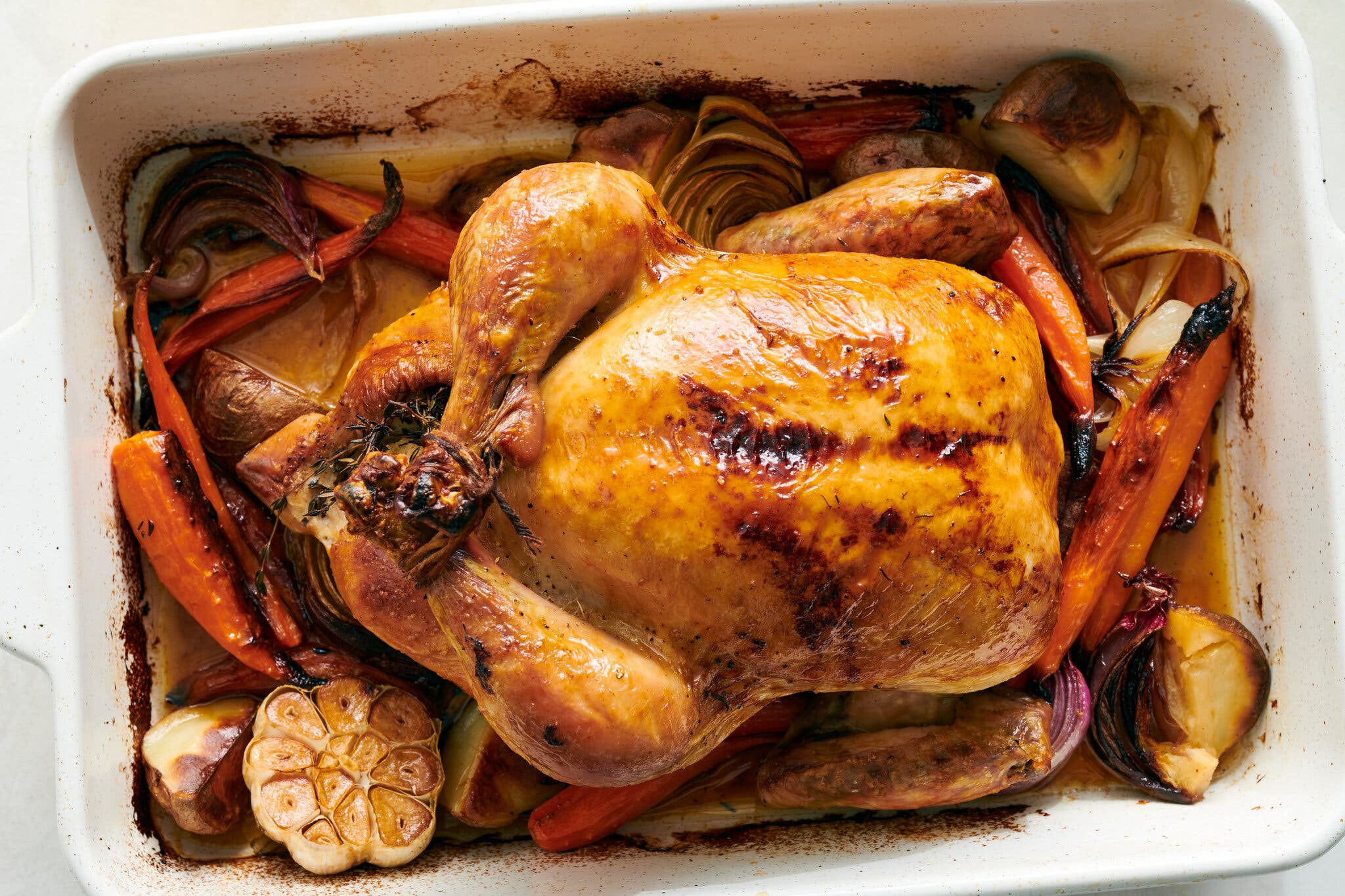 An overhead shot of a roast chicken sitting atop a bed of vegetables in a white roasting pan.
