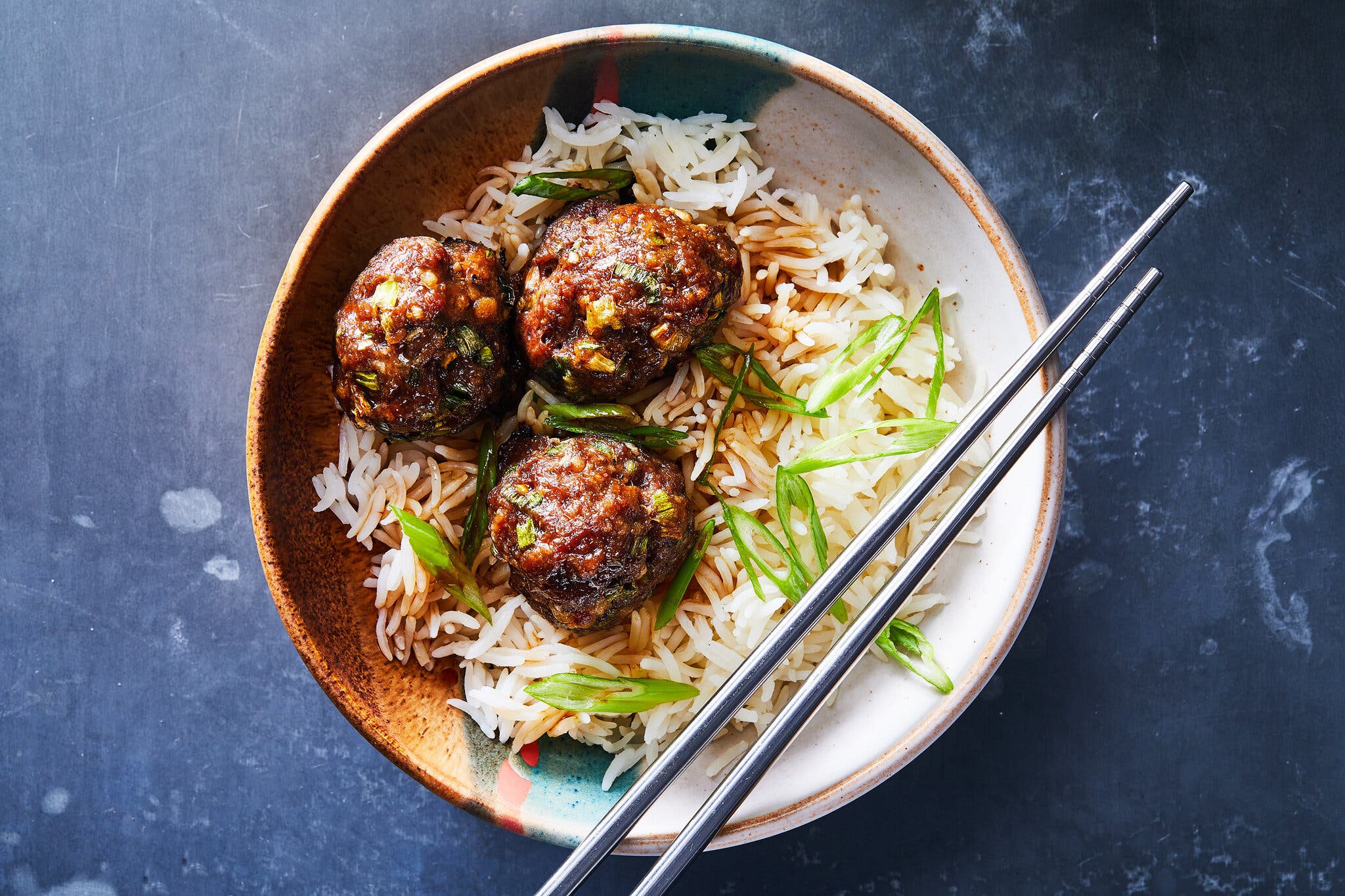 An overhead view of three dark brown meatballs topped with green onions, resting on a bed of white rice with a brown sauce; a pair of metal chopsticks lies across the speckled ceramic bowl.