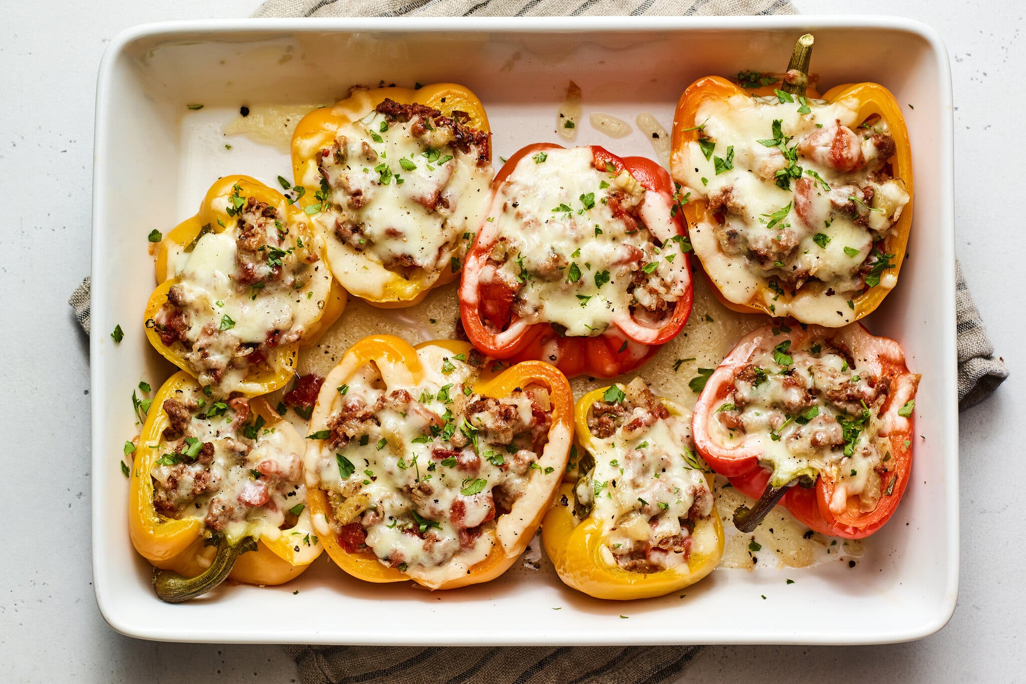 An overhead close-up of baked stuffed peppers, including five yellow and three red halves, filled with a brown meat mixture and covered with bubbly melted white cheese and green parsley.
