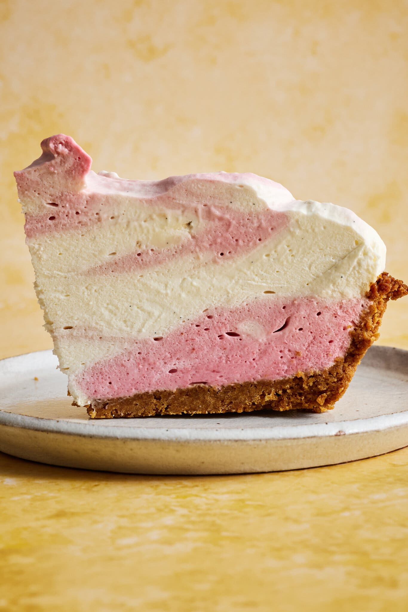A side view of a tall slice of pie with swirled white and pink mouse on a white plate against a yellow background.
