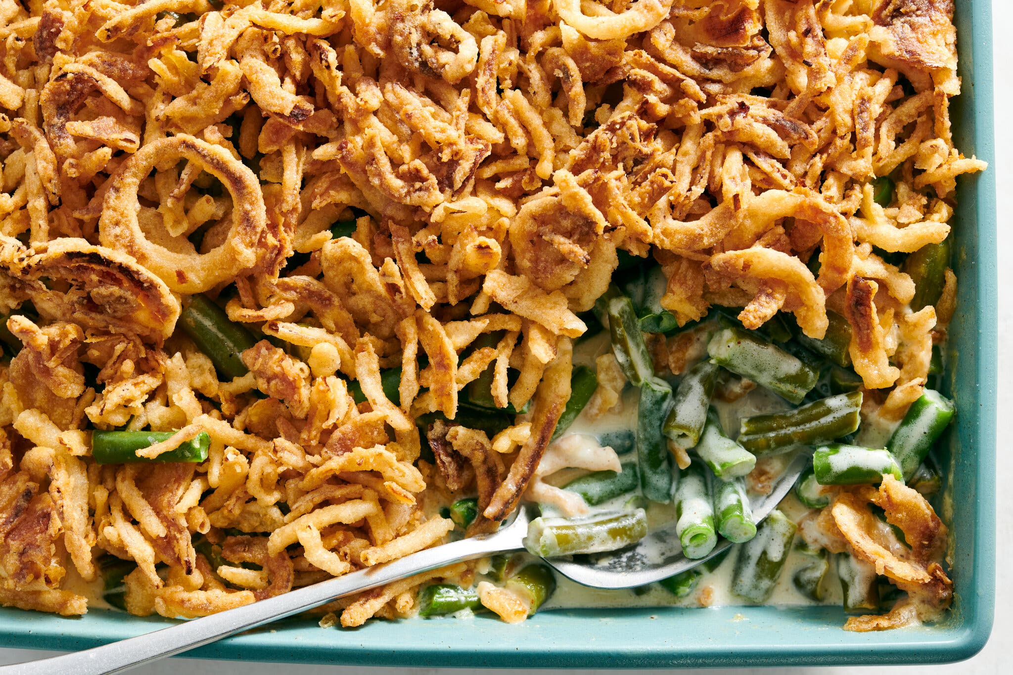 A green bean casserole fills a blue dish, with creamy green beans under golden fried onions. A silver spoon sits in one corner.