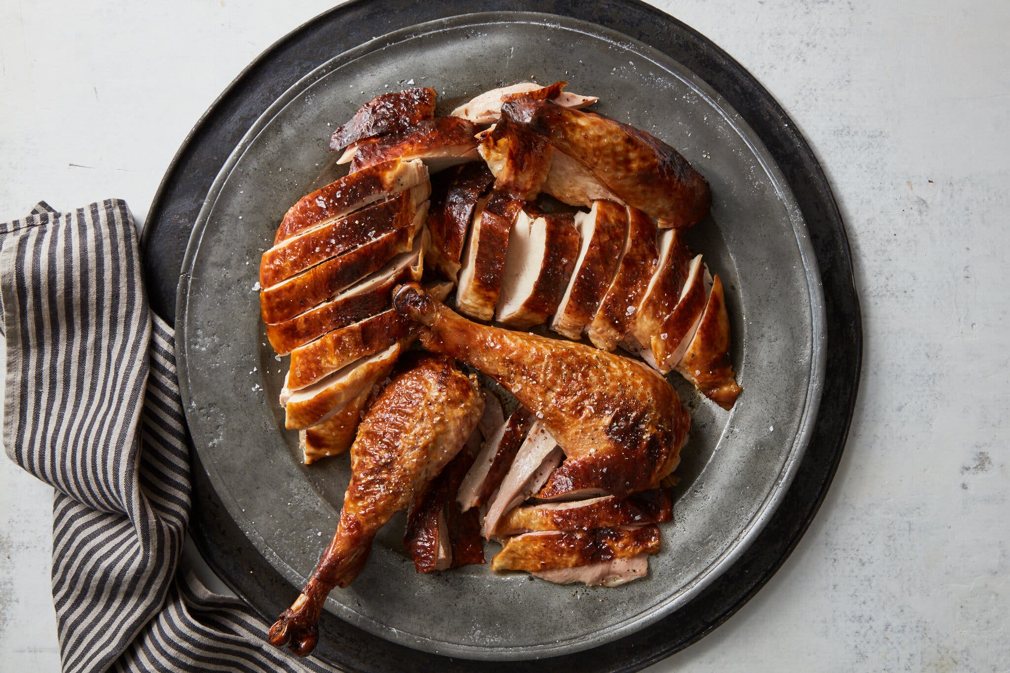 Carved roasted turkey, with golden-brown skin and sprinkles of flaky salt, sits on a dark metal platter.